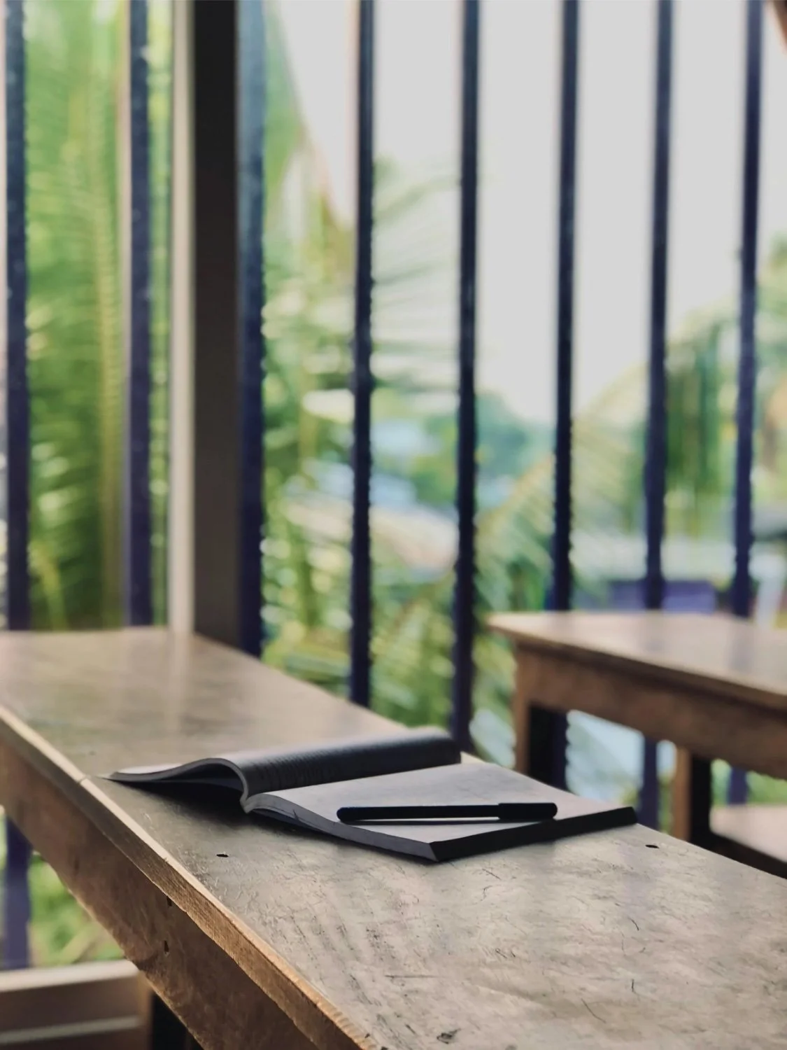 An open notebook and a black pen placed on a wooden table on a balcony with green foliage in the background.
