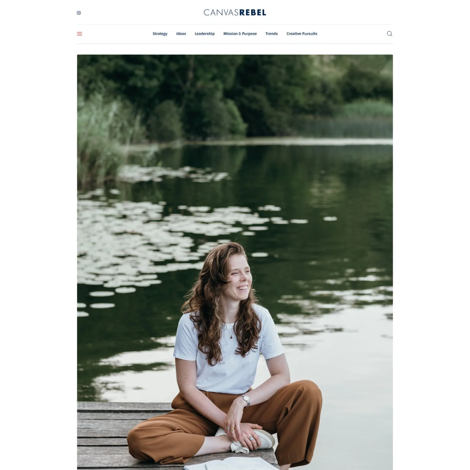A young woman with long, wavy brown hair sitting on a wooden dock by a lake, smiling and looking to the side, with trees and water lilies in the background.