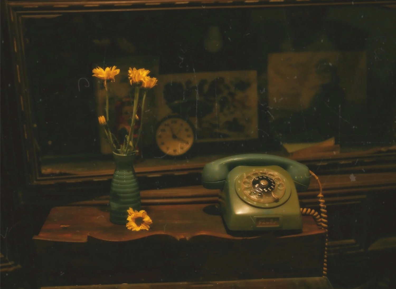 An old green rotary telephone sitting on a wooden table, next to a green vase containing yellow flowers, with a clock and vintage pictures in the background.