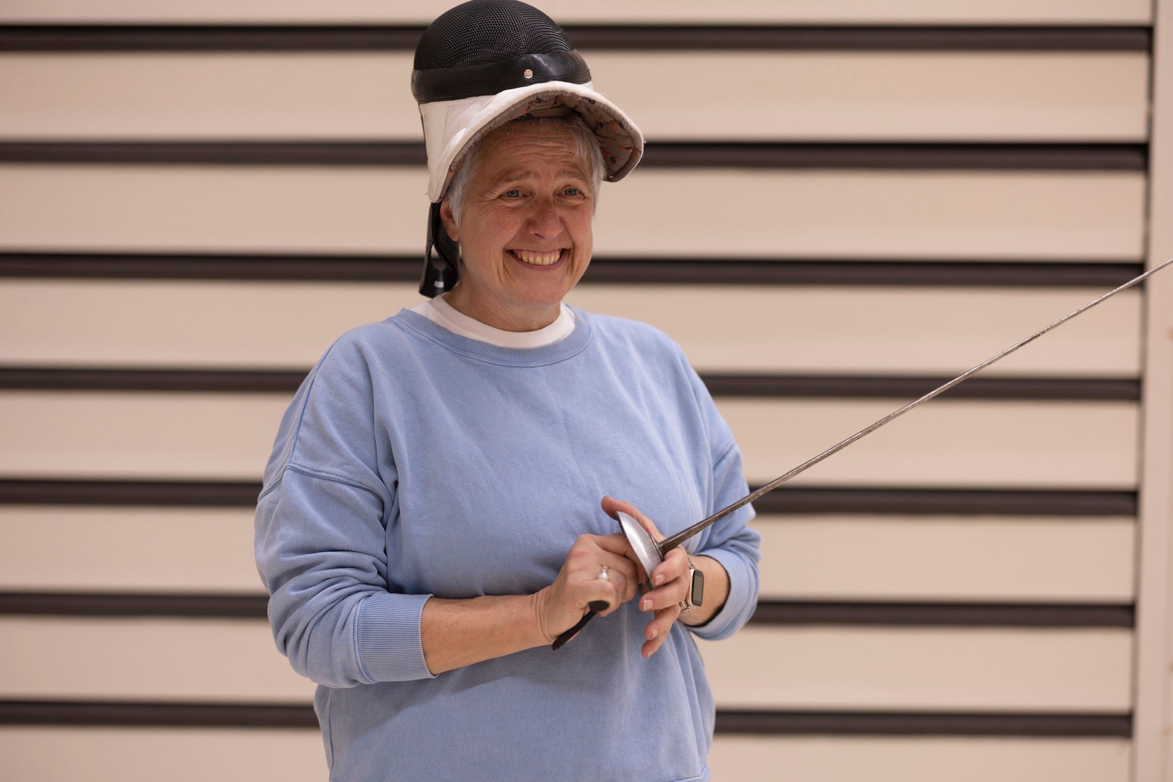 Person in blue sweatshirt holding a fencing foil, wearing a fencing mask on their head, smiling.