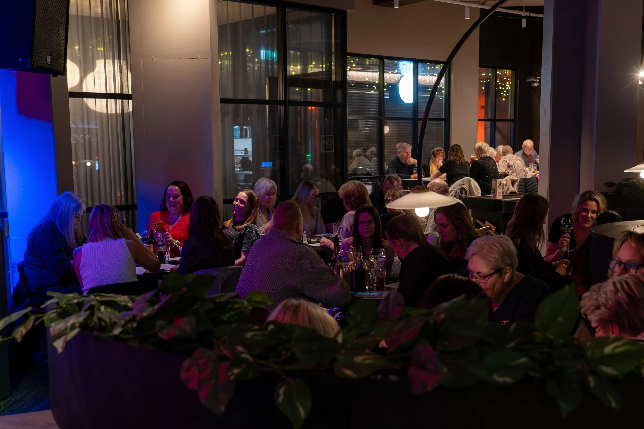 Interiørbilde av et folksomt restaurant eller kafé med mennesker som sosialiserer, spiser og drikker. Mørk belysning gir en koselig atmosfære med flere bord fylt med gjester.