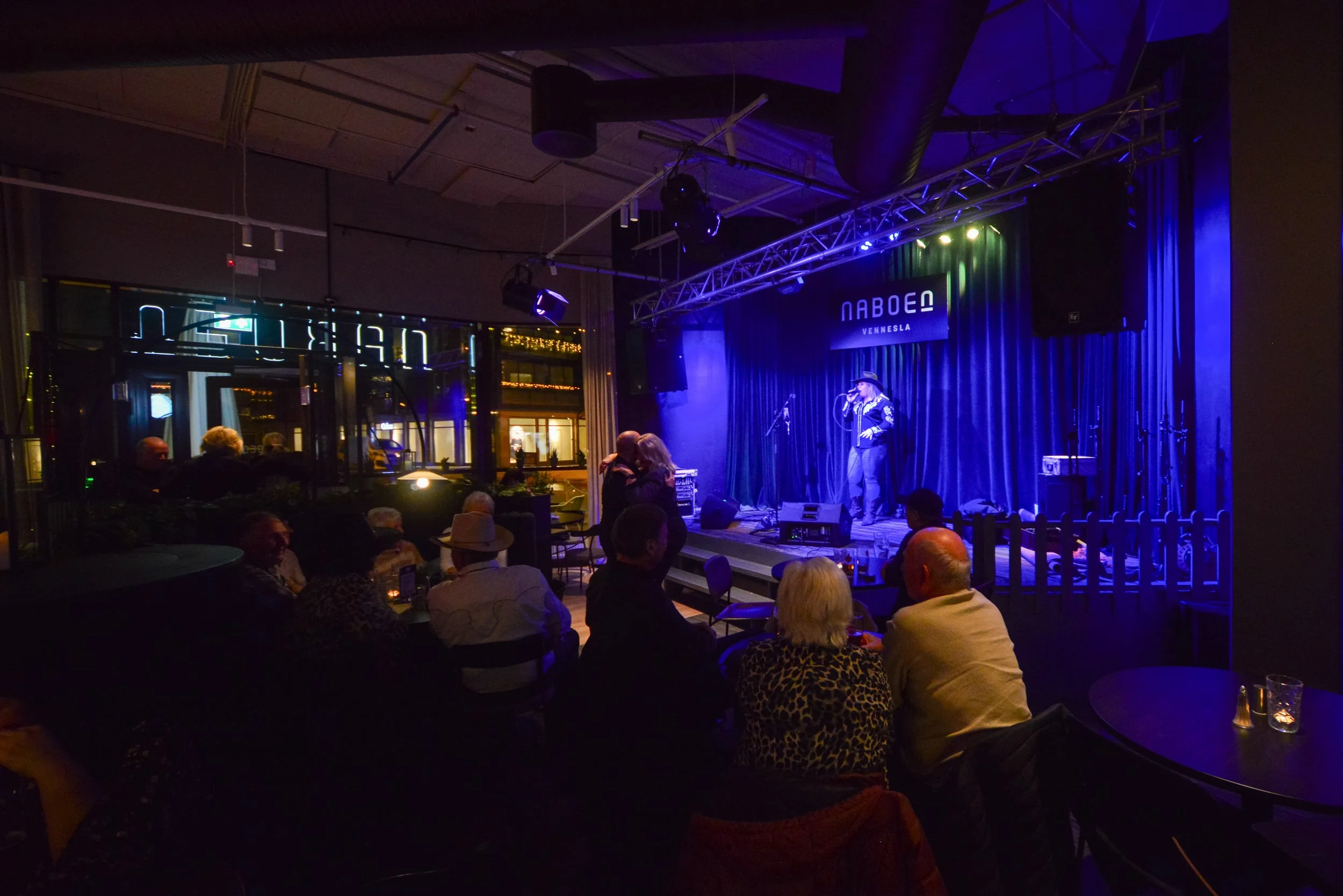 Innendørs scene med mennesker som sitter og ser på en opptrenden artist på en liten scene med blå bakgrunn. Det er mørkt og stemningsfullt, med blå og lilla belysning.