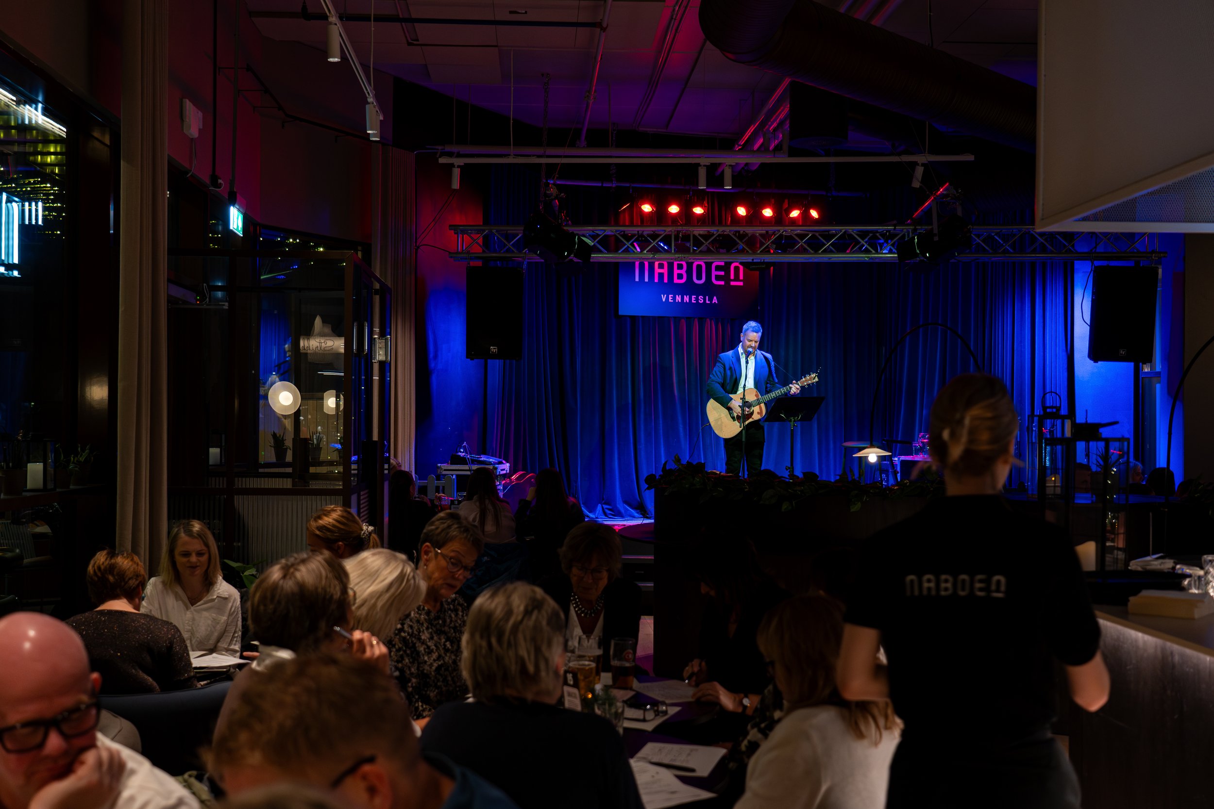 En innendørs konsertscene med en mann som spiller akustisk gitar og synger, omgitt av mørkeblå gardiner og rød lyssetting. Publikum sitter rundt bord, og en servitør i svart uniform med teksten 'NABOEN' på ryggen serverer.