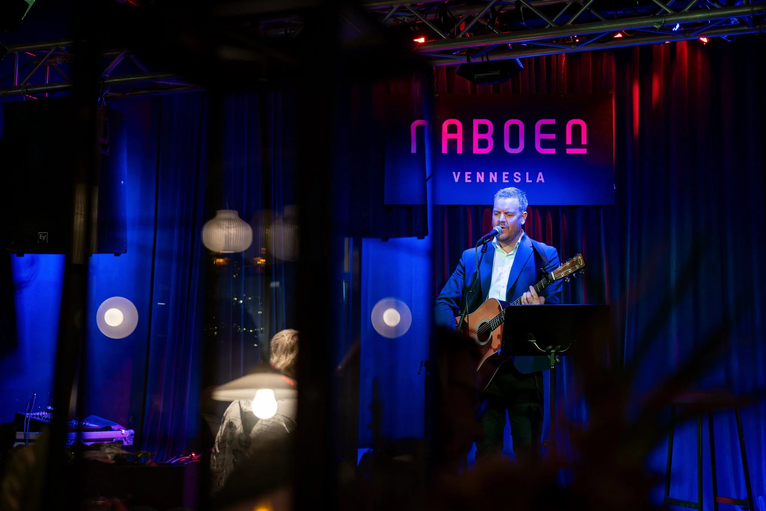 En mann spiller gitar og synger inn i mikrofon på en scen med mørke blå og røde gardiner, foran en neonlyssign som sier 'NABO' og 'VENNSLA'.