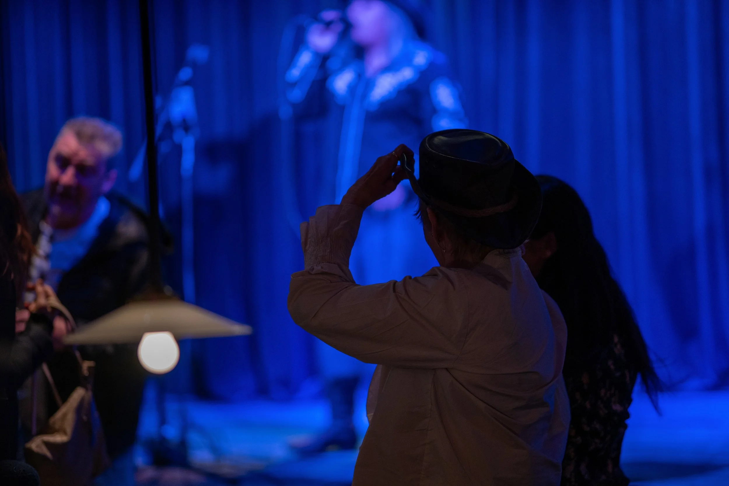 En person iført hatt tar bilde av en scene med folk og en kvinne som synger på en scenekledd i blå lys.