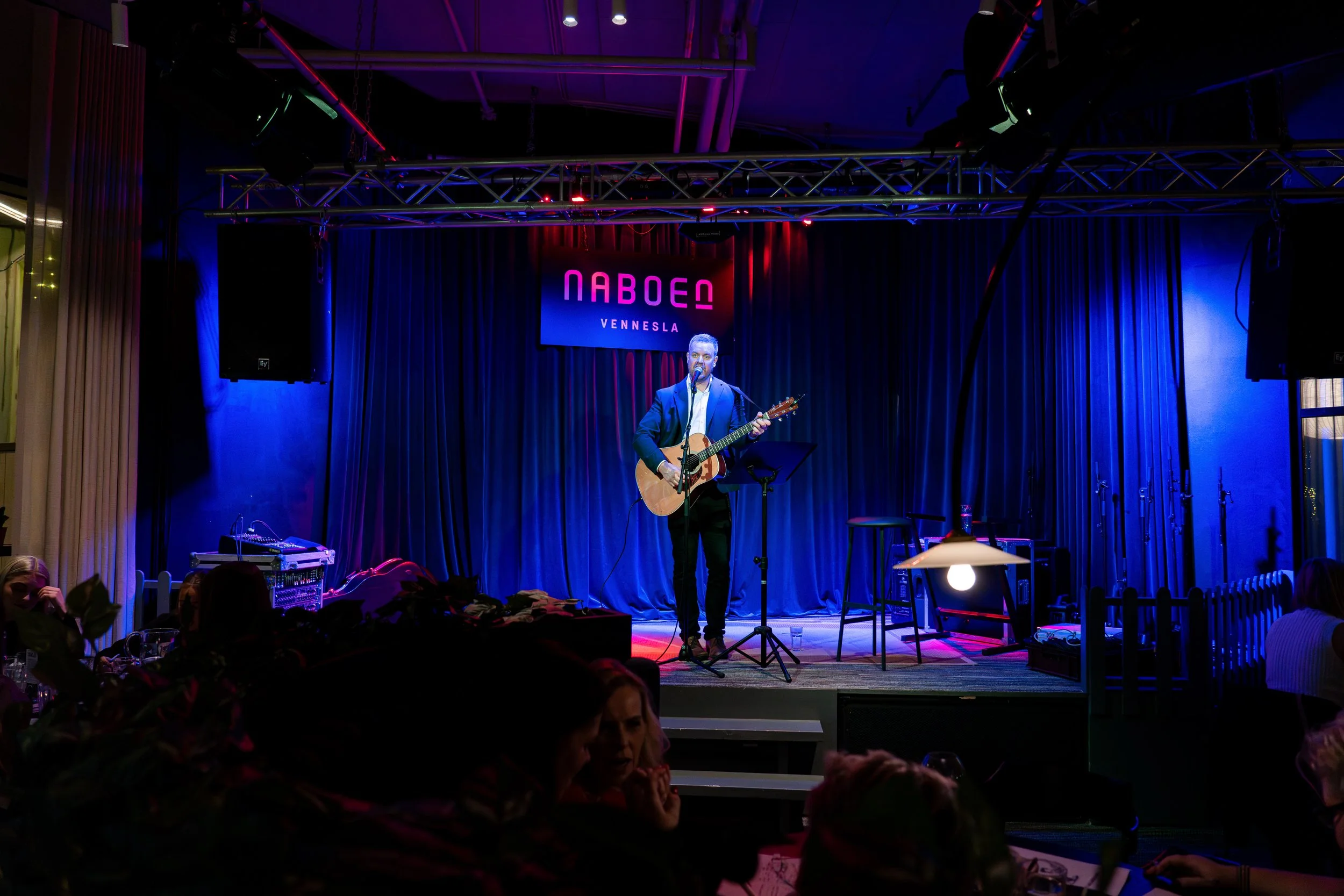 En musiker spiller akustisk gitar på en scene med blå og lilla lys, bak en mørkeblå gardin, under et skilt som sier 'NABOEN VENNESLA'.