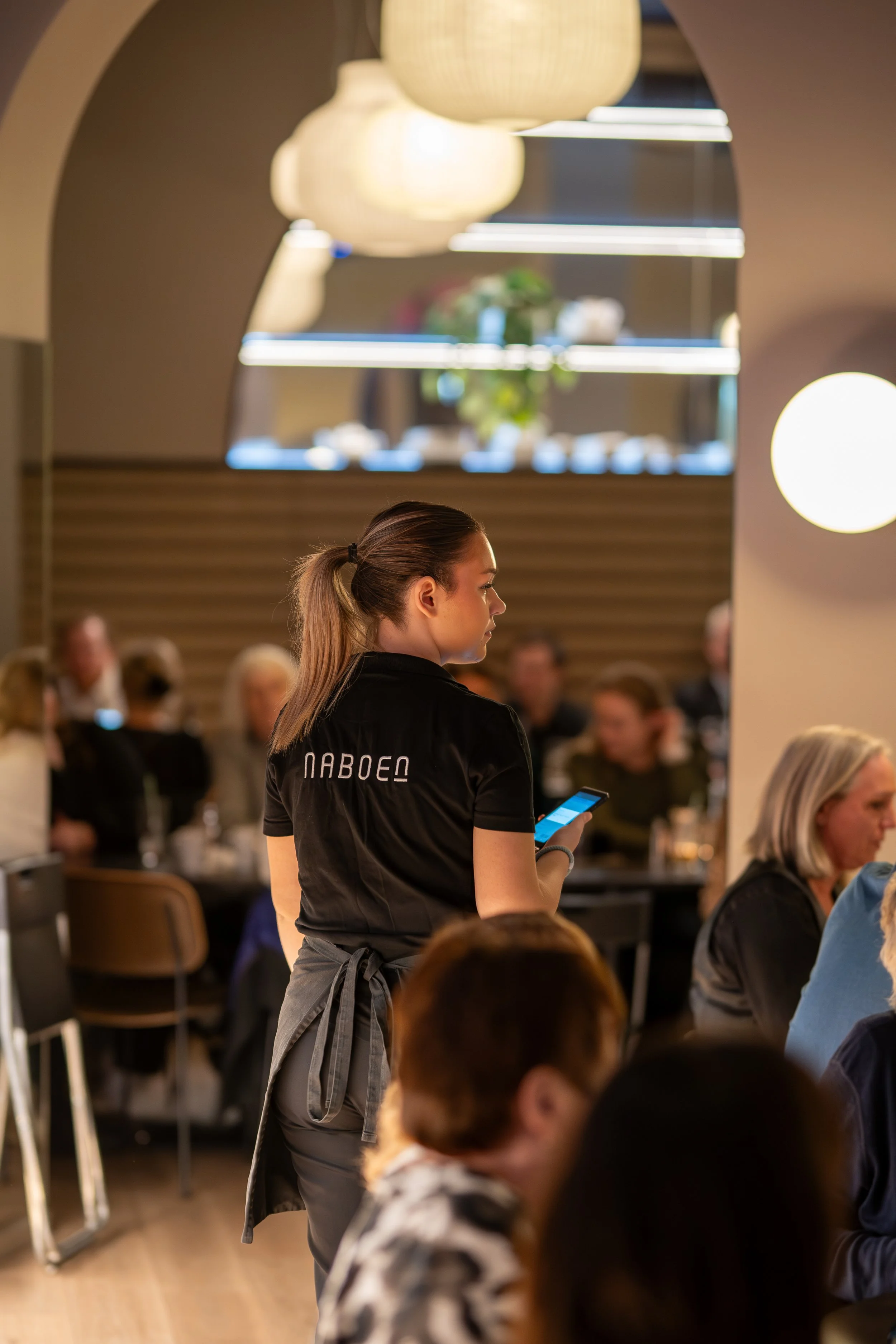 En servitør i svartuniform står i en restaurant, ser på telefonen sin, med mange gjester rundt henne.