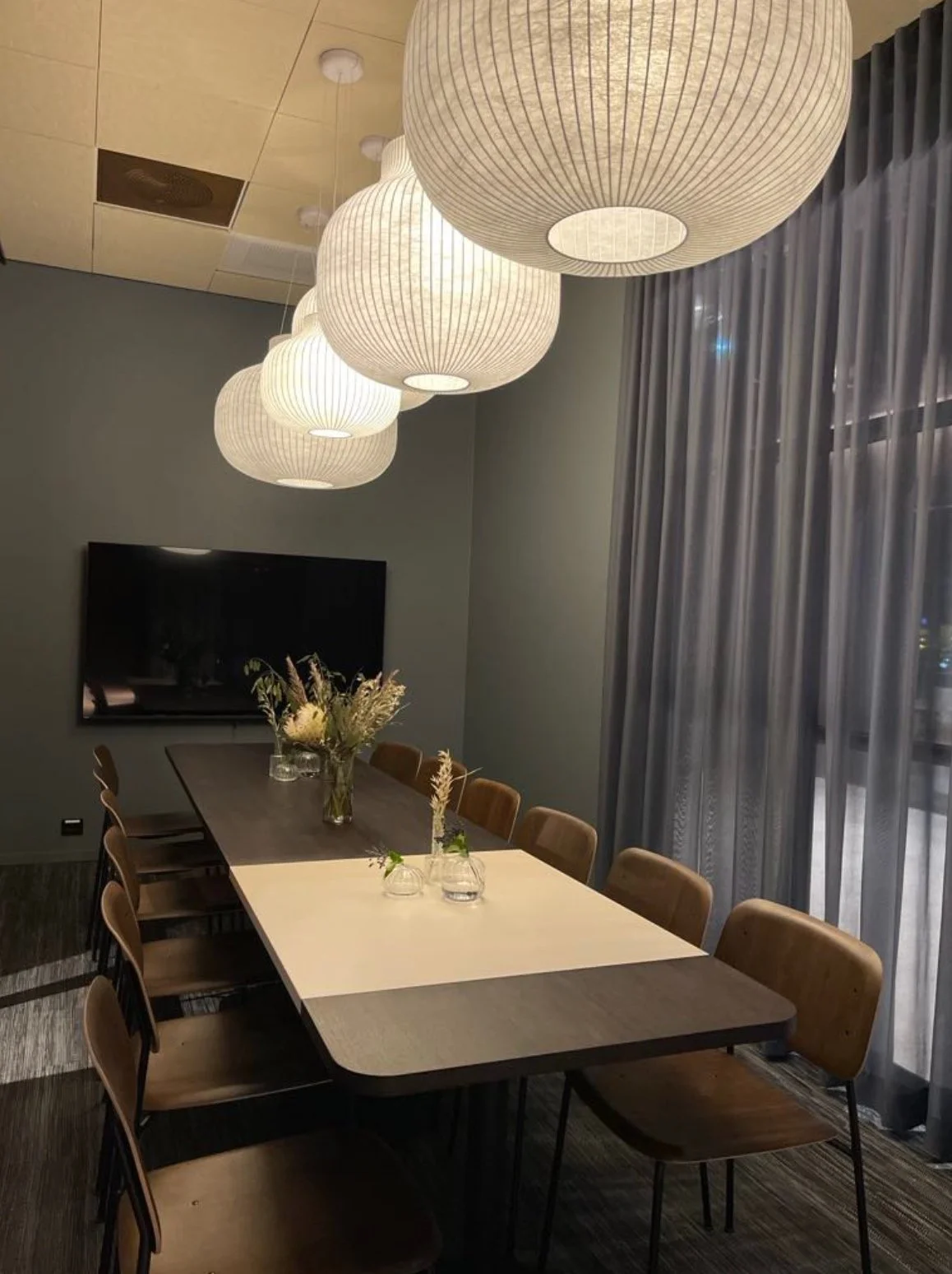 Et møtebord med åtte tremøblerte stoler rundt, plassert i et rom med store vinduer og sorte gardiner. Tre vaser med blomster står på bordet, og det henger fem store, runde papirlamper i taket.