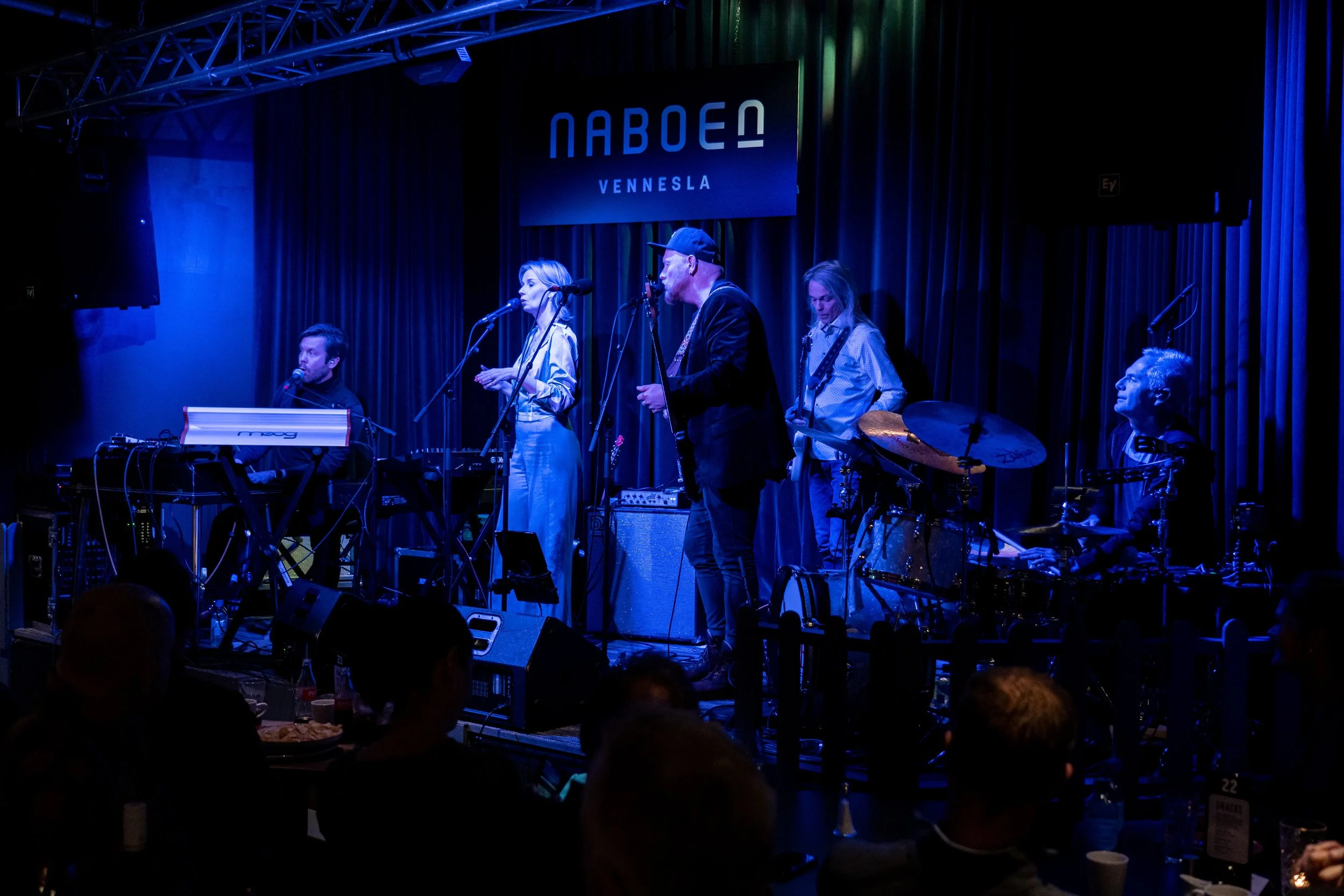 Band spiller på scene med blå lys, med logo «NABOEN Vennesla» i bakgrunnen, i en innendørs konsertsal.