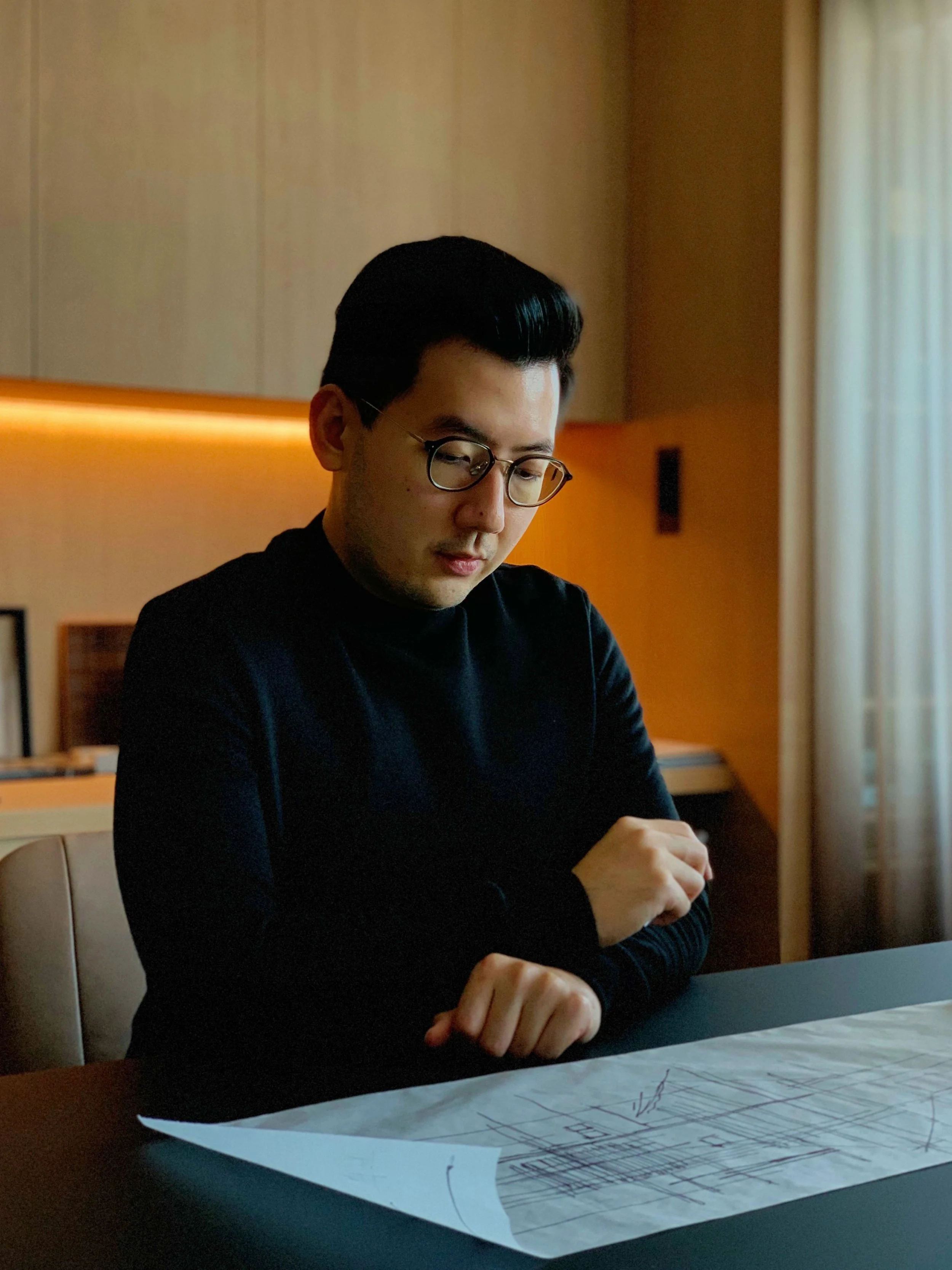 A man with dark hair and glasses sitting at a table with architectural plans, in a room with warm lighting and light-colored curtains.