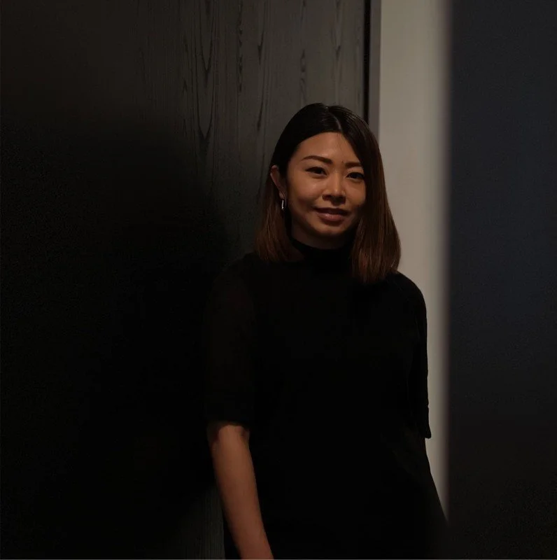 A woman with shoulder-length brown hair, wearing a black top, standing against a dark wall.