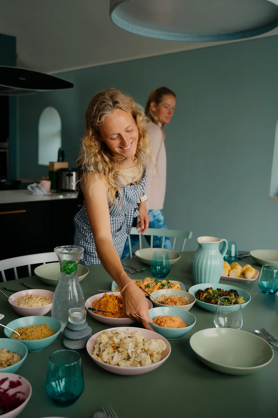 Zwei Frauen beim Abendessen in einem gemütlichen Raum. Eine Frau lächelt und serviert sich Essen von einem Tisch mit verschiedenen Speisen, während die andere im Hintergrund steht.
