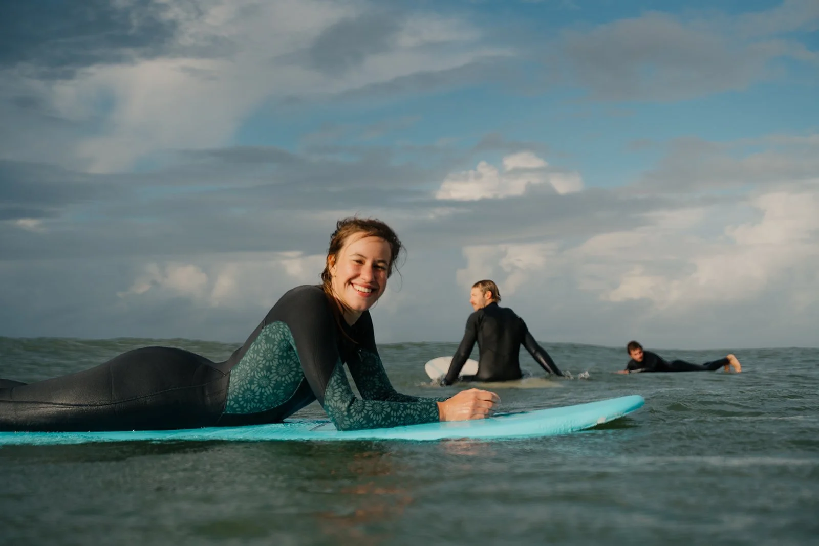 Drei Menschen beim Surfen im Meer, die Frau im Vordergrund lächelt in die Kamera, die anderen zwei Männer sind weiter im Wasser, die eine Person liegt auf dem Bauch, die andere sitzt auf einem Surfbrett.