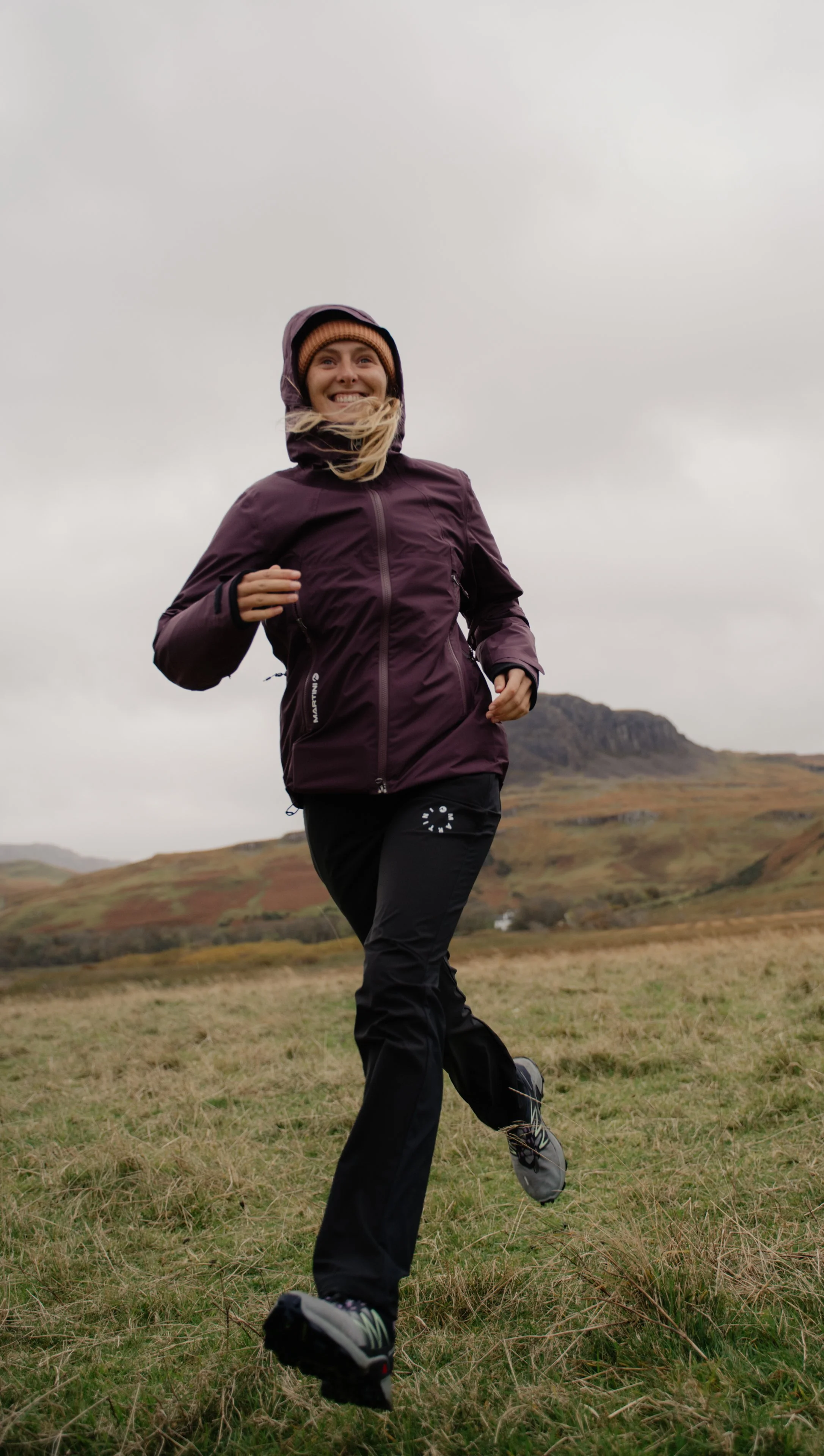 Eine lächelnde Frau läuft in einer offenen, grünen Landschaft bei bewölktem Himmel, trägt eine dunkelviolette Jacke, schwarze Hose und Laufschuhe.