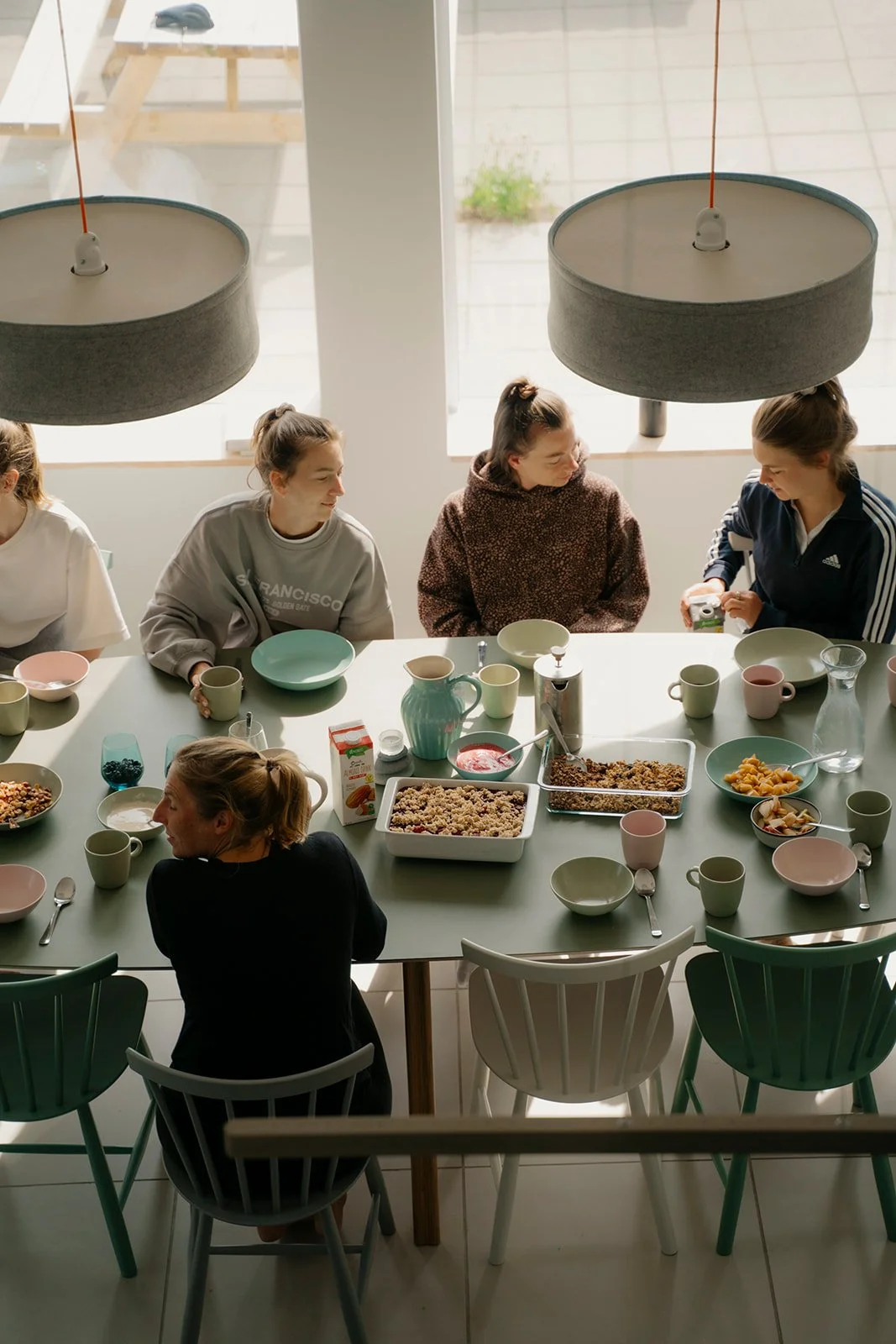 Eine Gruppe von Frauen sitzend an einem Esstisch mit Frühstück. Der Tisch ist mit Schalen, Tassen, einer Keksschale, einer Milchpackung und anderen Frühstücksartikeln gedeckt. Die Frauen unterhalten sich, während sie frühstücken.