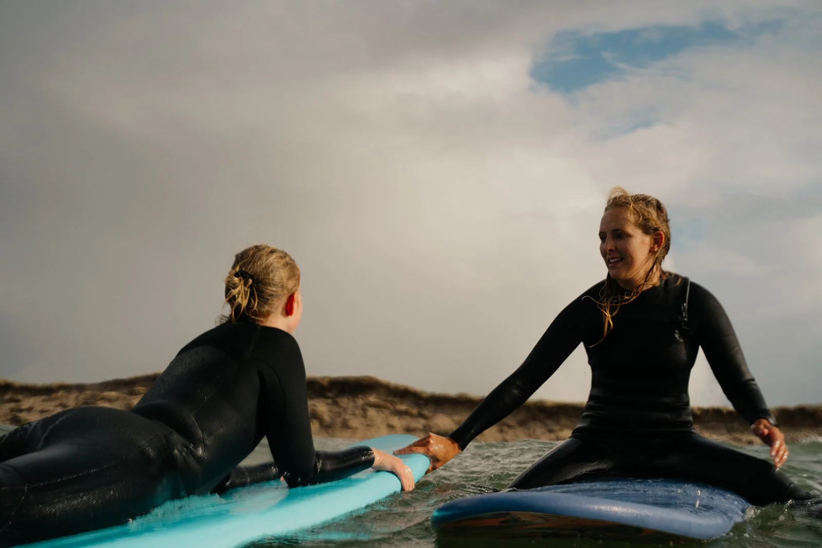 Zwei Frauen beim Surfen im Wasser, eine reicht der anderen die Hand, beide tragen Neoprenanzüge, am Strand mit Himmel im Hintergrund.