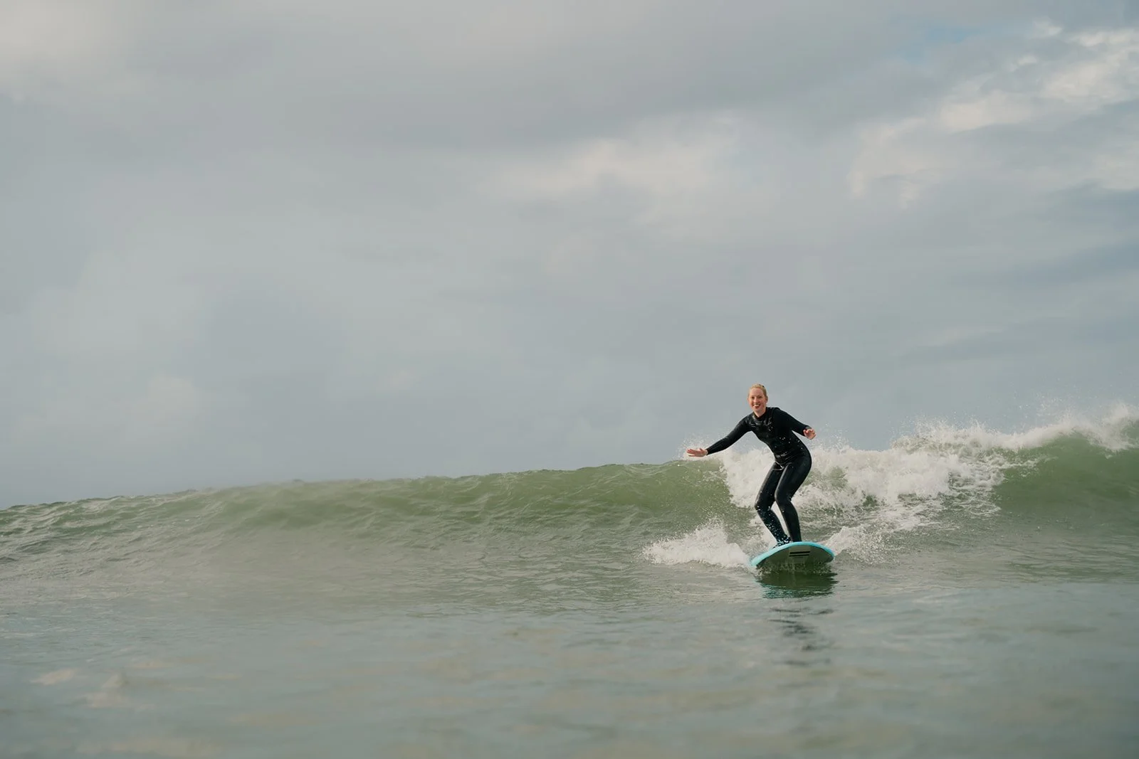Eine Frau beim Surfen auf einer Welle im Meer, trägt einen schwarzen Neoprenanzug, mit einem breiten Lächeln auf dem Gesicht.