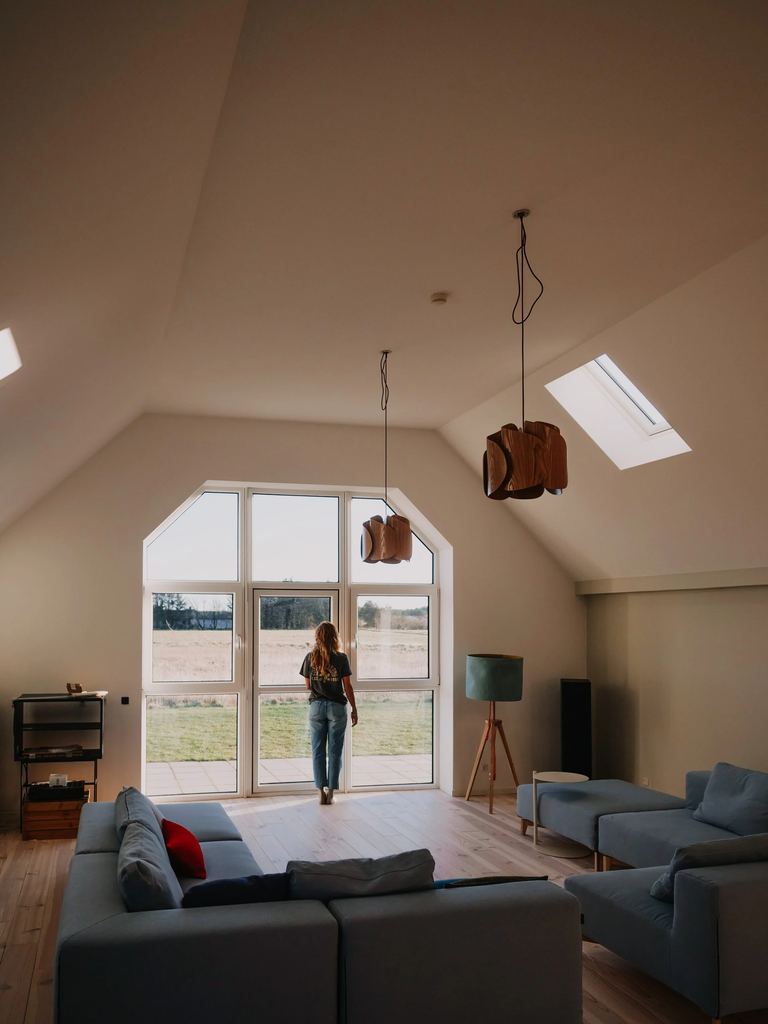 Ein modernes Wohnzimmer mit hohen Decken, großen Fenstern und einer Frau, die nach draußen schaut. Es gibt Sofas, einen Bodenlampenständer und zwei hängende Leuchten aus Holz. Das Zimmer ist hell und minimalistisch gestaltet.