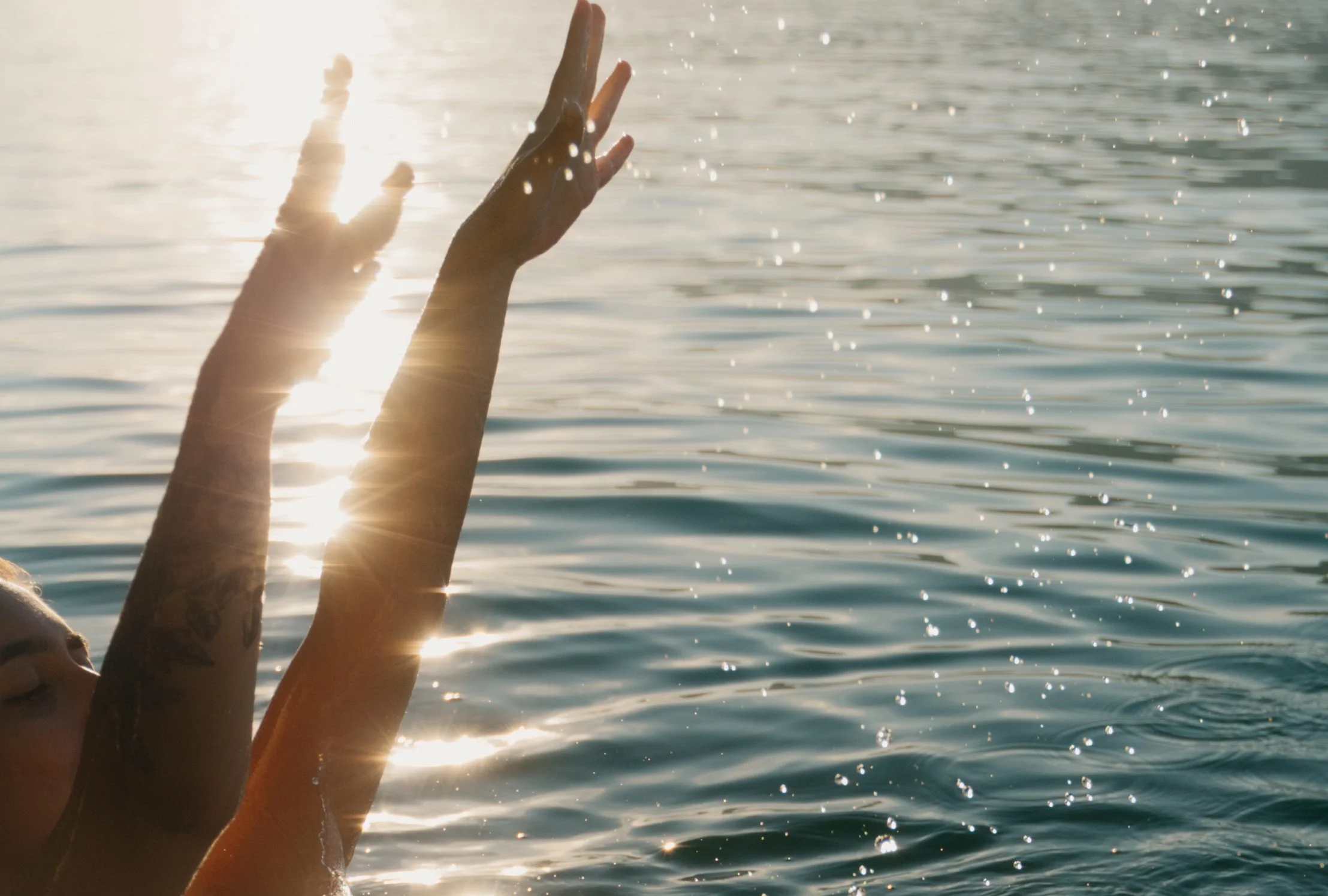 Nähe eines Sees, eine Person mit Tattoos hebt ihre Hände im Wasser, Sonne spiegelt auf der Wasseroberfläche, Wasser spritzt umher.