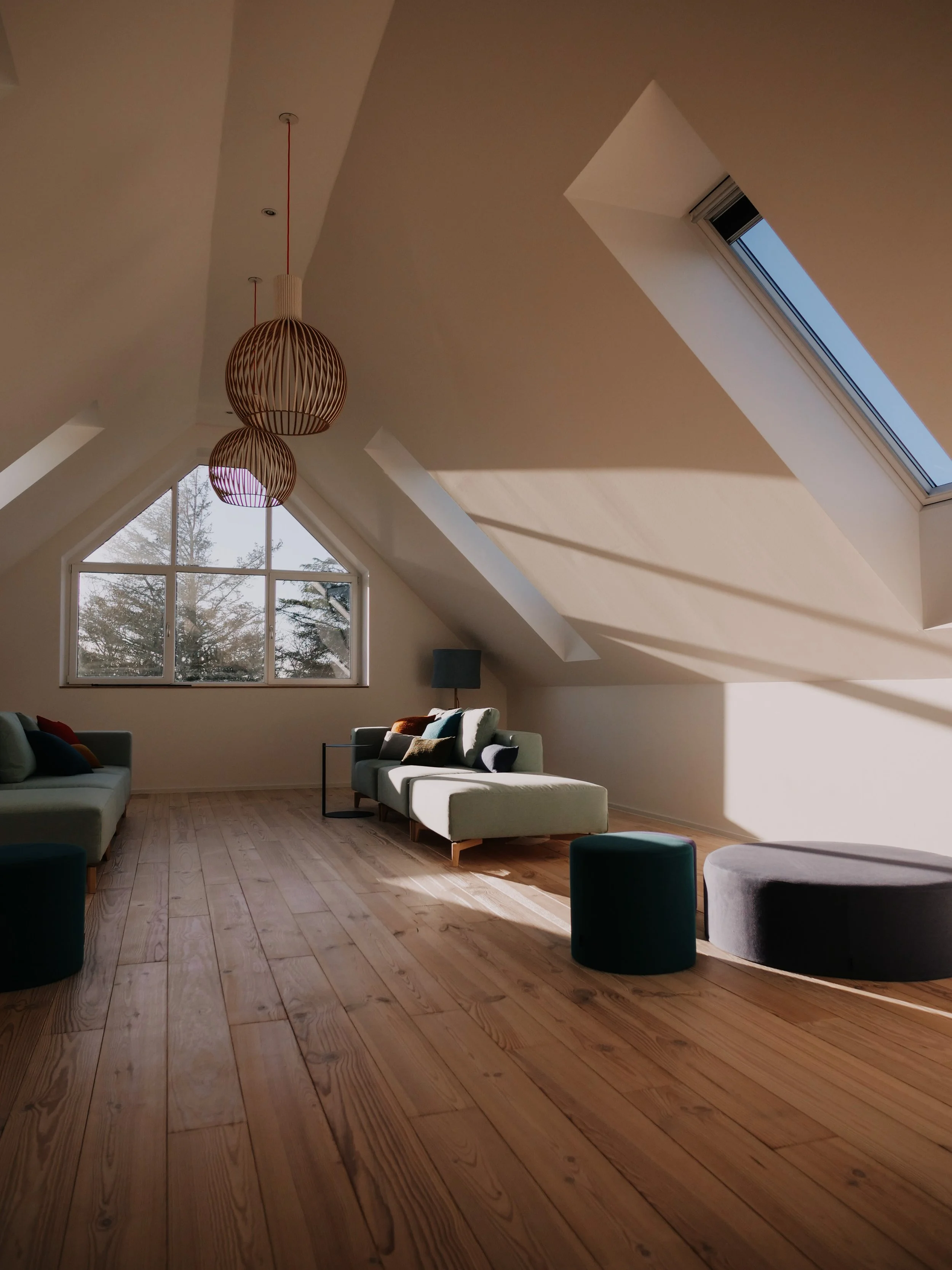 Ein modernes Wohnzimmer im Dachgeschoss mit Holzboden, großen Fenstern und einem schrägen Dach, das durch die Fenster natürlichen Lichteinfall hat.