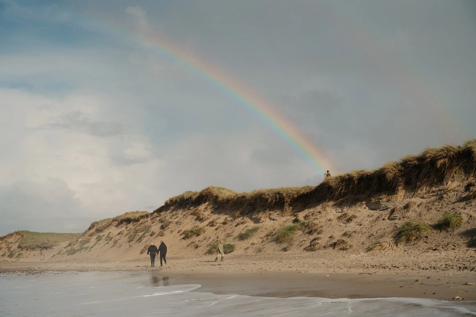 Strand mit Düne, Spaziergänger, Regenbogen am Himmel, bewölkter Himmel
