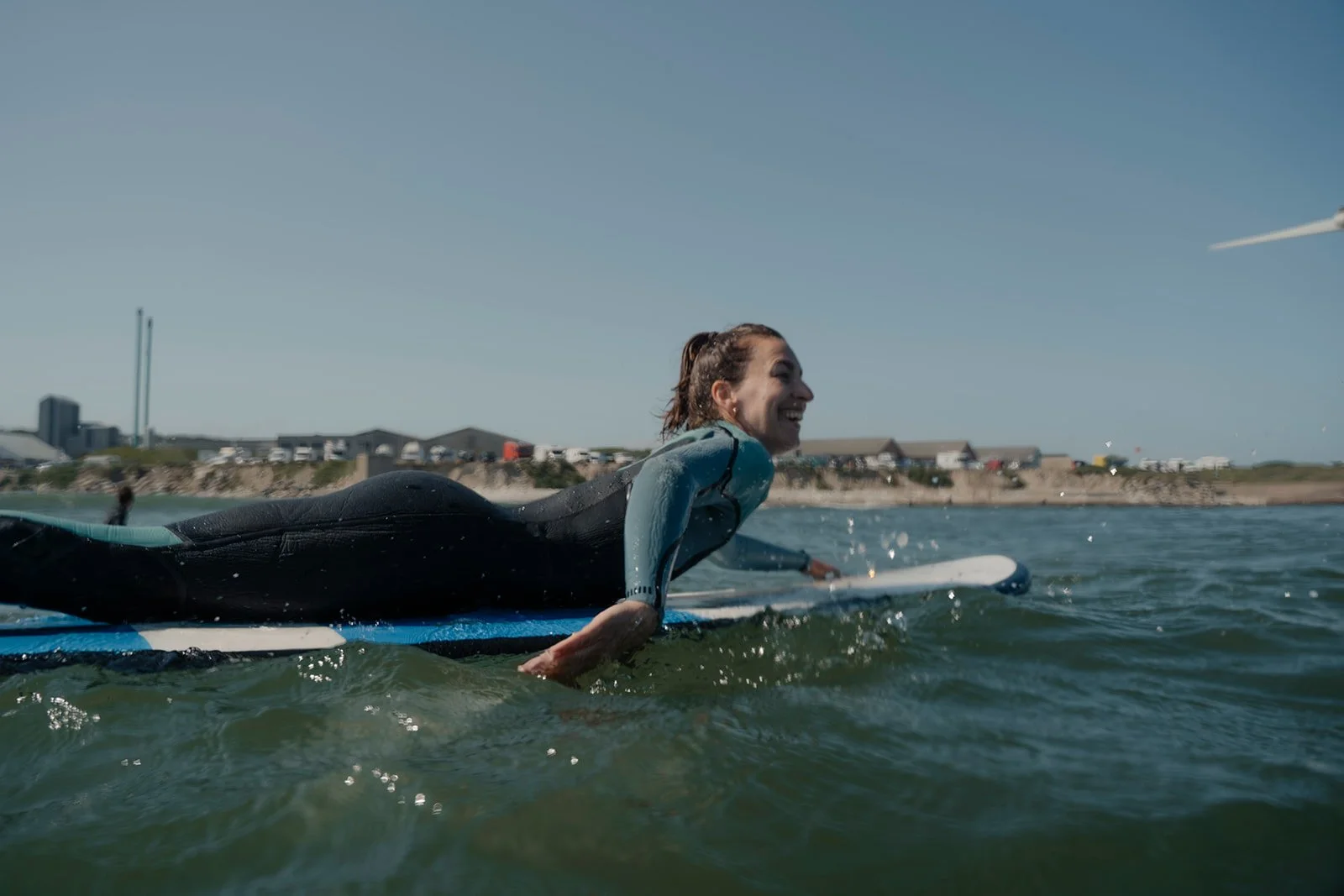 Frau beim Surfen im Meer, lächelnd, im Neoprenanzug, mit Küste und Gebäuden im Hintergrund.