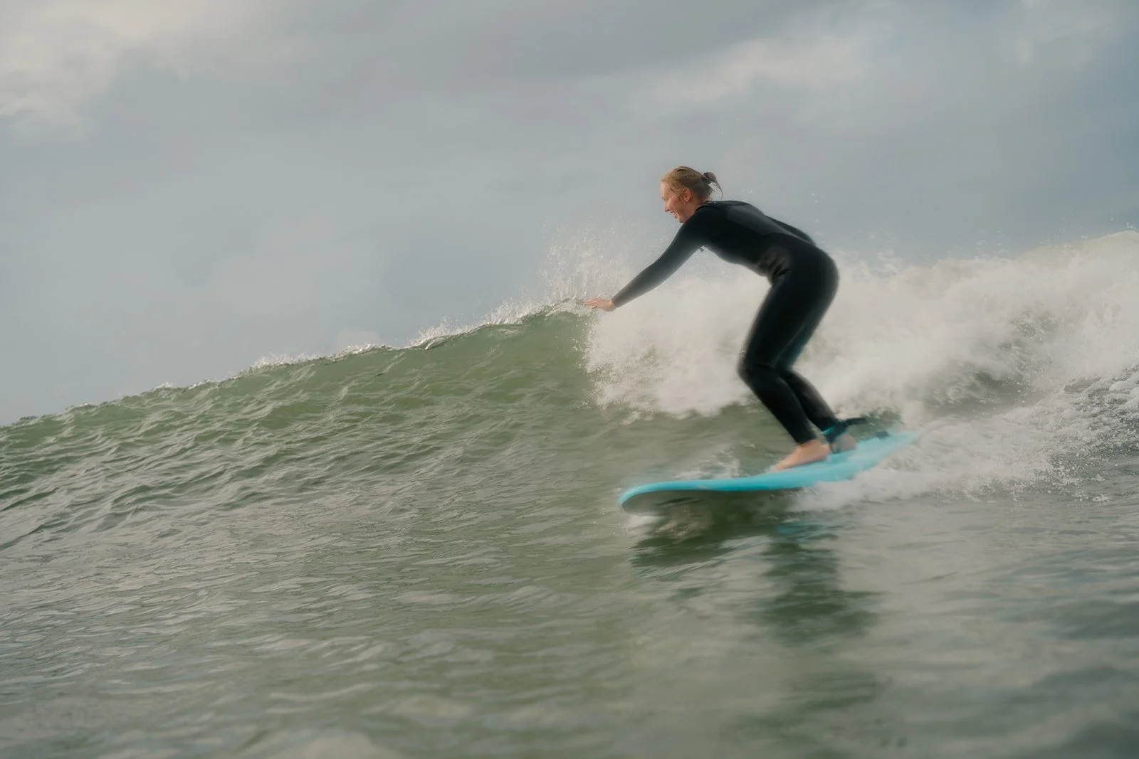 Eine Frau surft auf einer Meereswelle, trägt einen schwarzen Neoprenanzug und hat die Haare zu einem Knoten zusammengebunden.
