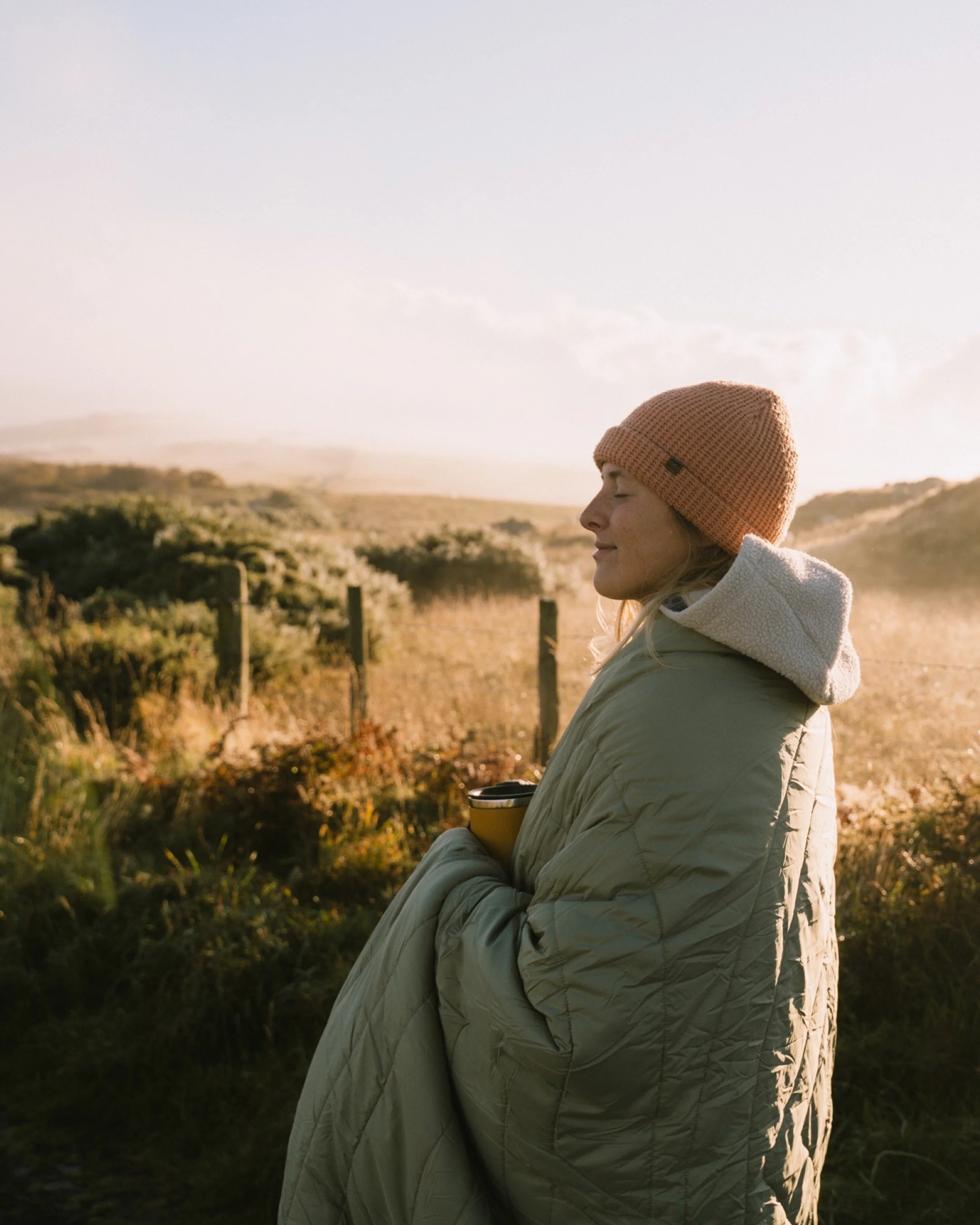 Frau in warmer Kleidung steht entspannt im Freien, hält einen Becher und genießt den Sonnenaufgang oder Sonnenuntergang in einer ländlichen Umgebung.