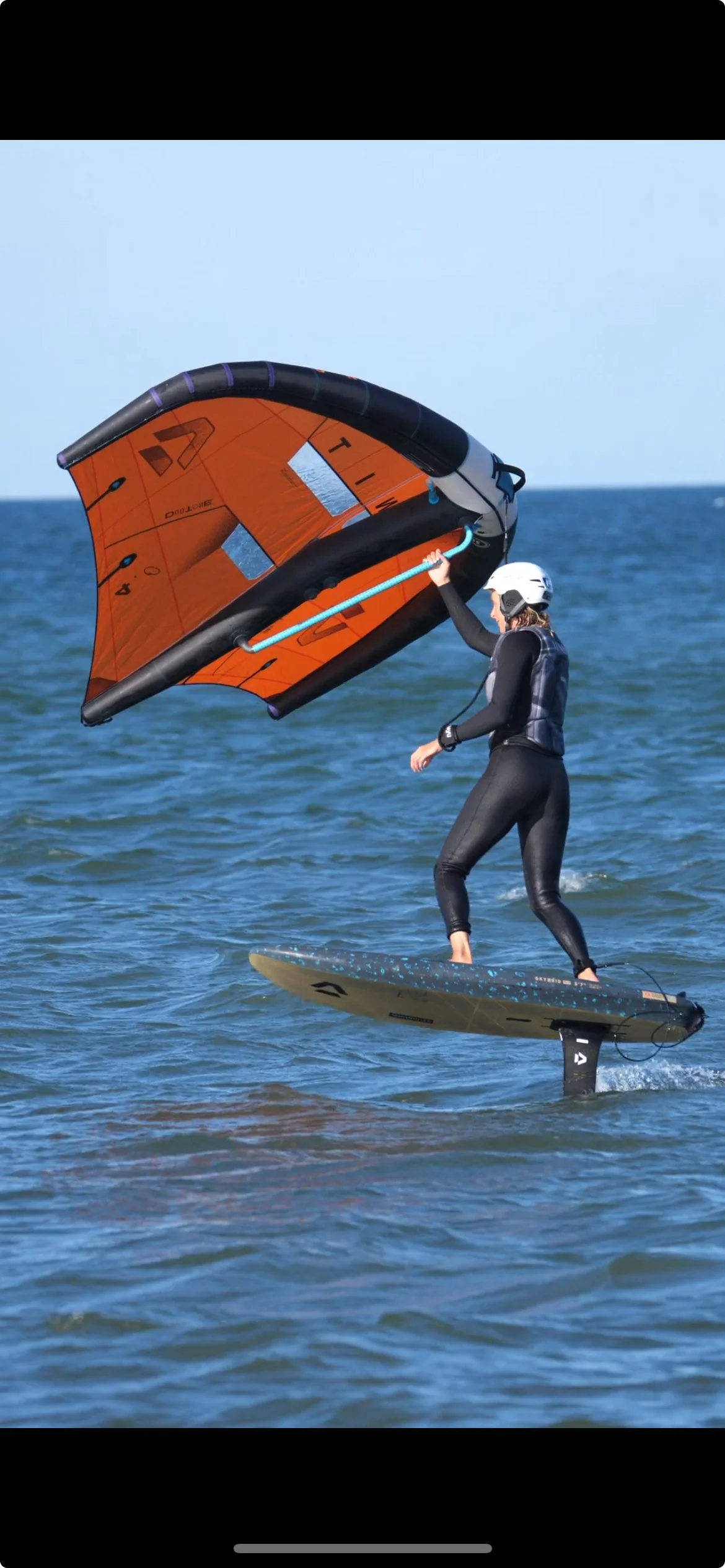Eine Person steht auf einem Wasser-Surfbrett mit Flossen im Wasser, hebt ein orangefarbenes Segel, um Windsurfen zu betreiben, während sie einen weißen Helm trägt.