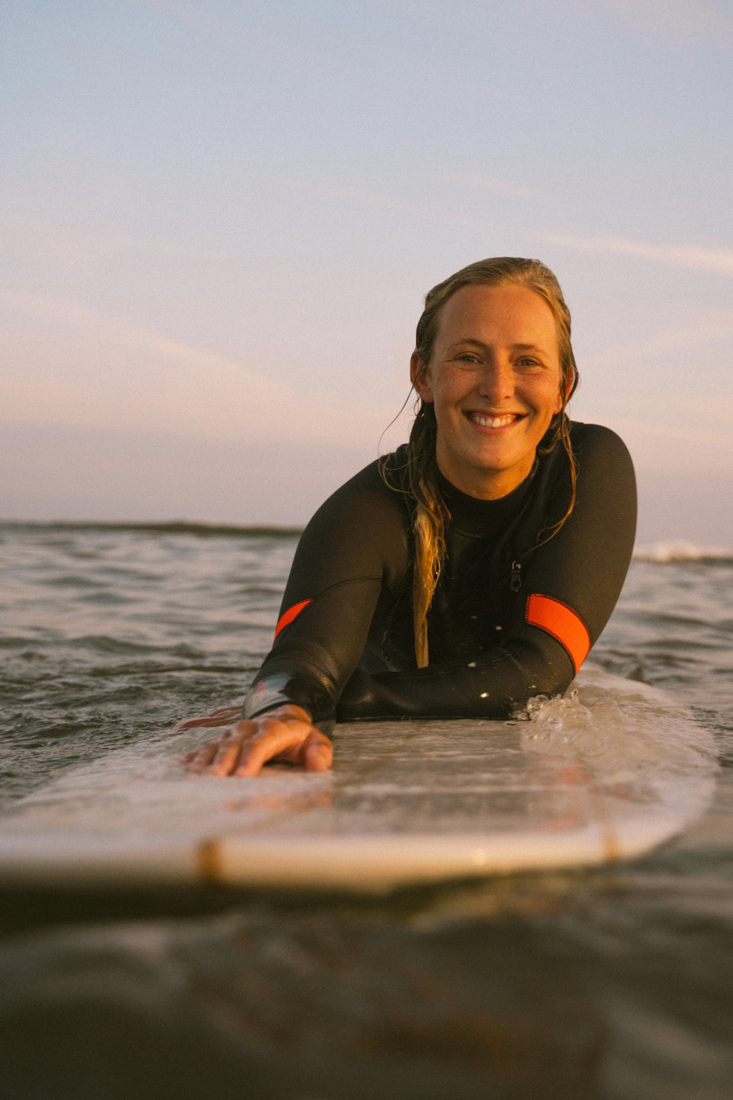 Junge Frau beim Surfen im Meer, lächelnd, bei Sonnenuntergang, trägt einen Neoprenanzug.