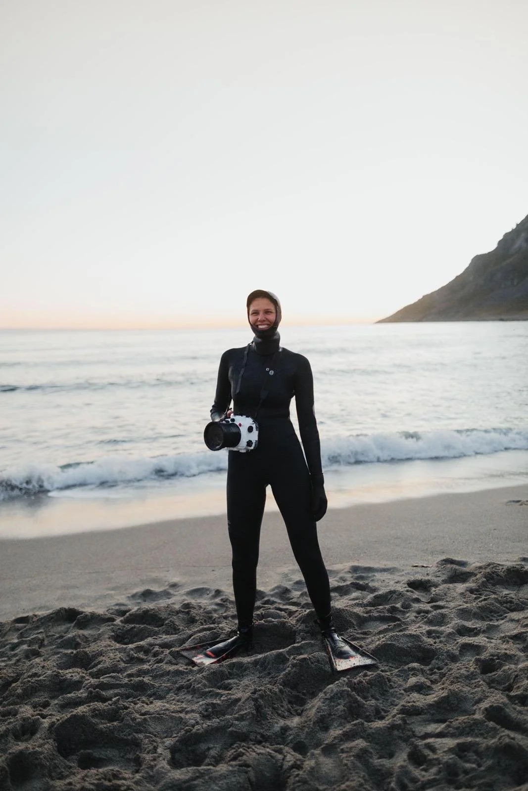 Eine junge Frau im Tauchanzug steht mit Flossen am Strand, hält eine Kamera und lächelt bei Sonnenuntergang mit Blick auf das Meer und eine Küste im Hintergrund.