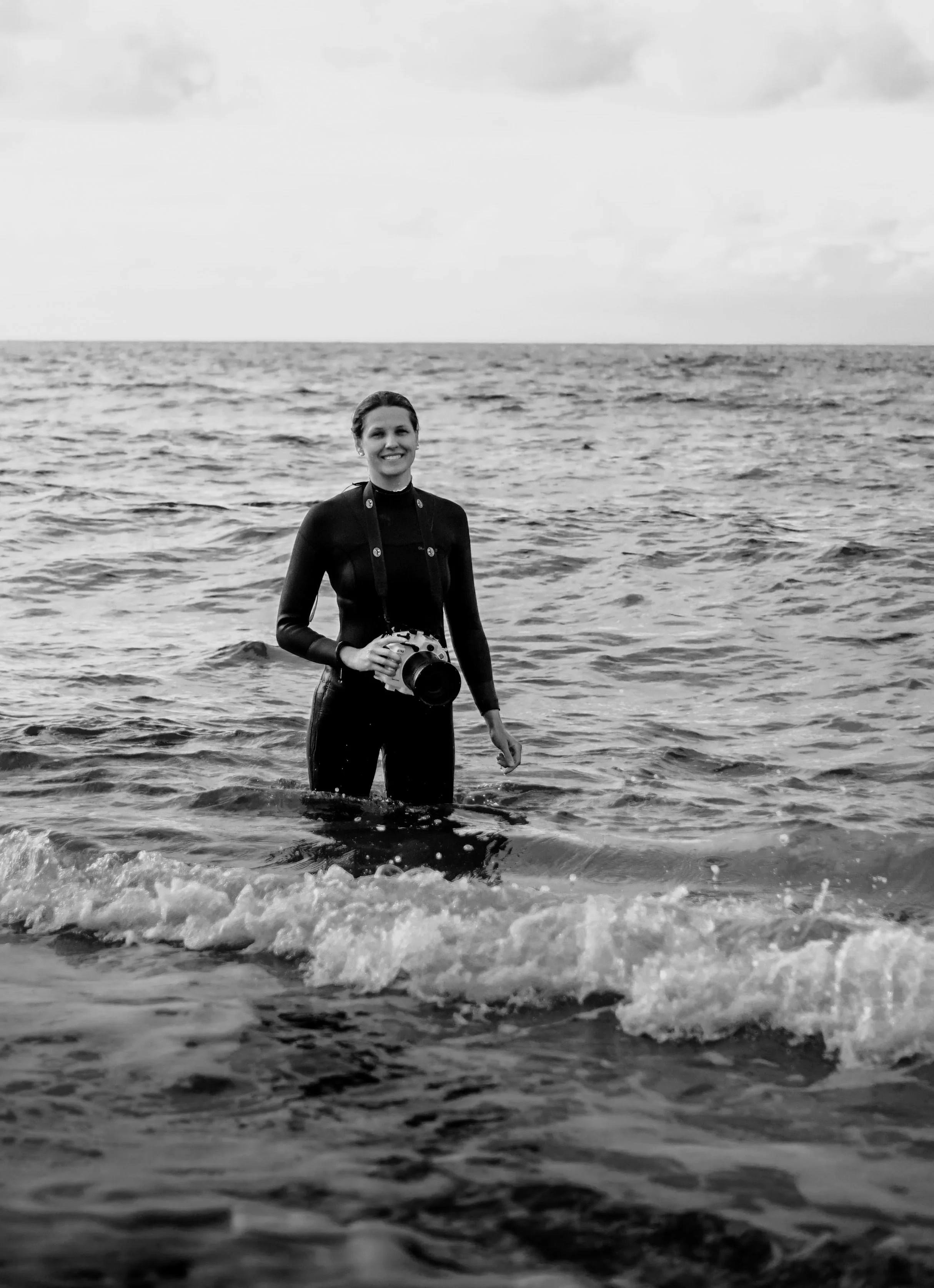Eine lächelnde Frau steht im Meer, hält eine Kamera und trägt einen Neoprenanzug.