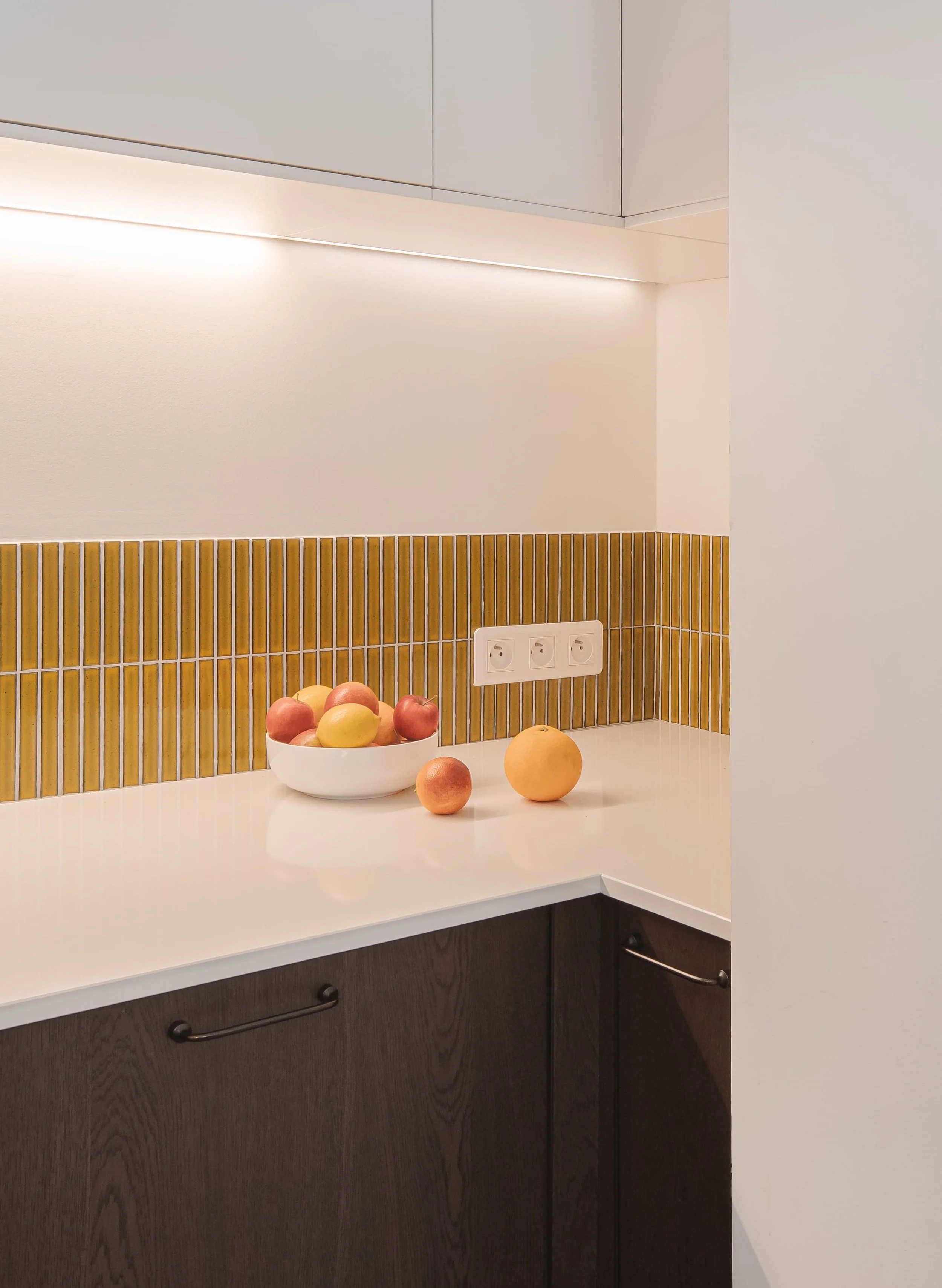 Une cuisine moderne avec un comptoir blanc, un dosseret en carrelage jaune, une prise électrique et un bol de fruits contenant des pommes, des pêches et des oranges.