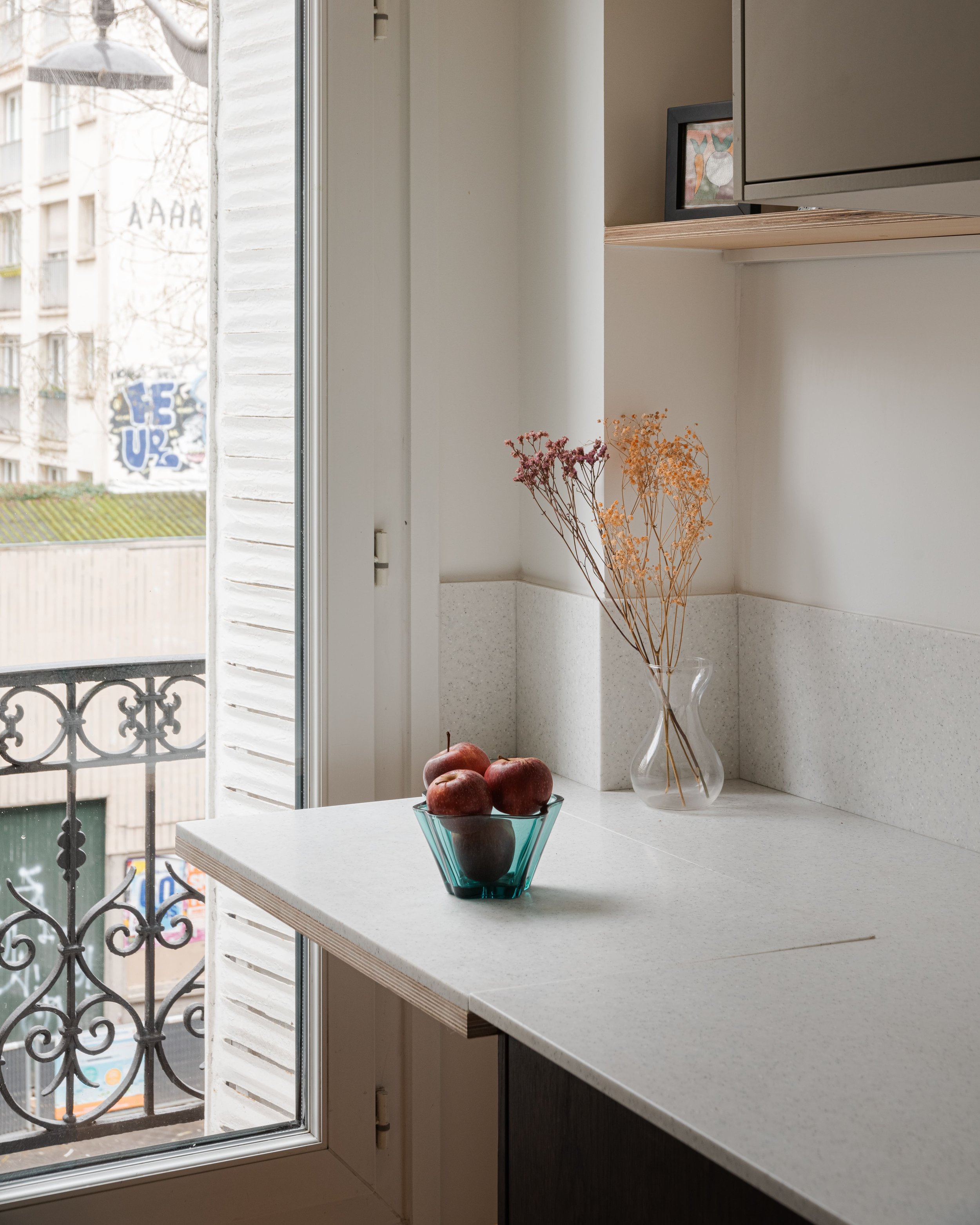 Une fenêtre donnant sur un balcon en fer forgé, avec une poignée de fruits rouges dans un bol en verre sur un comptoir de cuisine blanc, et un vase transparent avec des fleurs séchées orange et rouges.