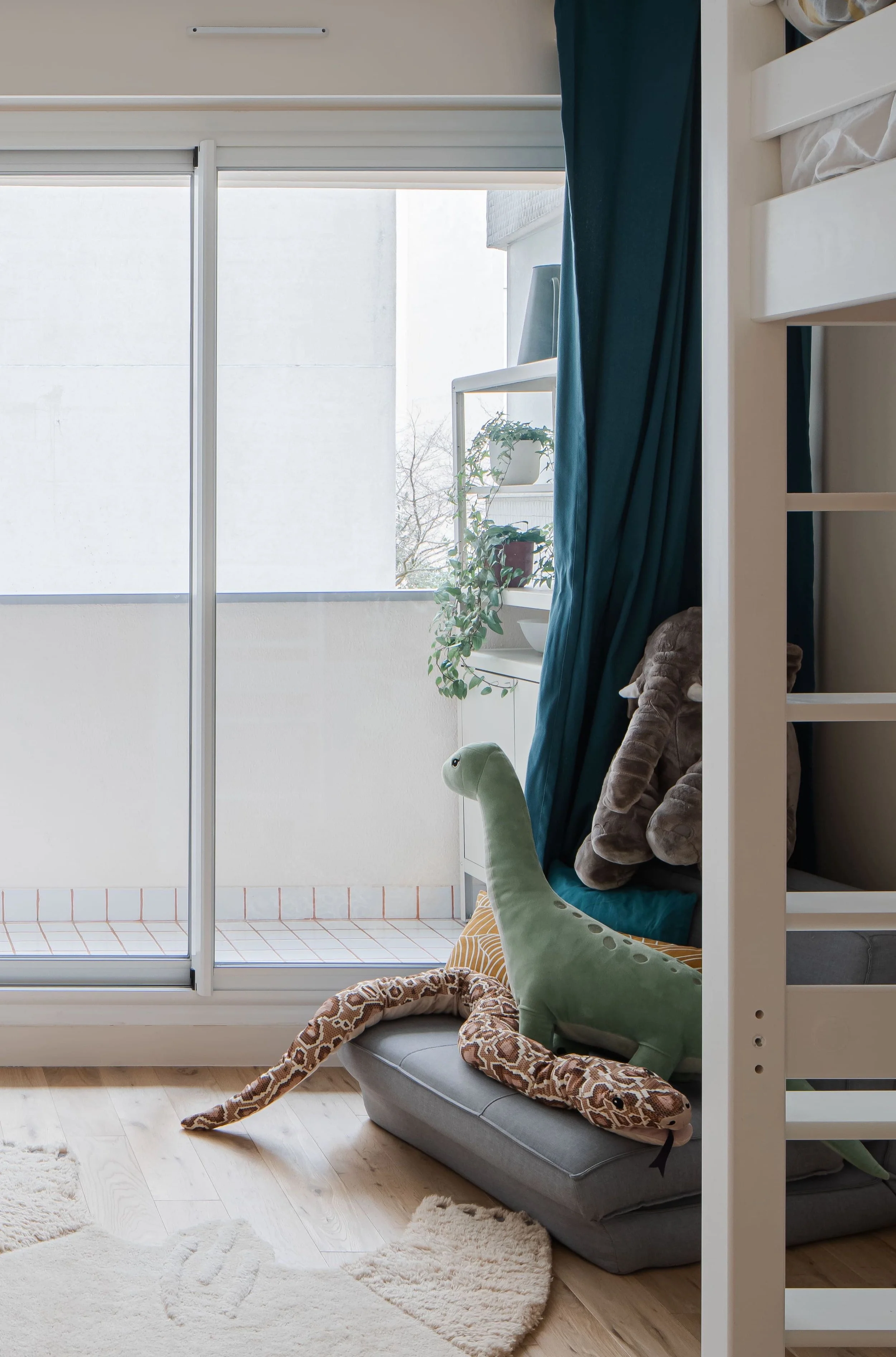 Chambre d'enfant avec un lit mezzanine blanc, rideaux bleus, un animal en peluche en forme de lézard vert, un éléphant en peluche, et un espace avec une porte-fenêtre donnant sur un balcon.