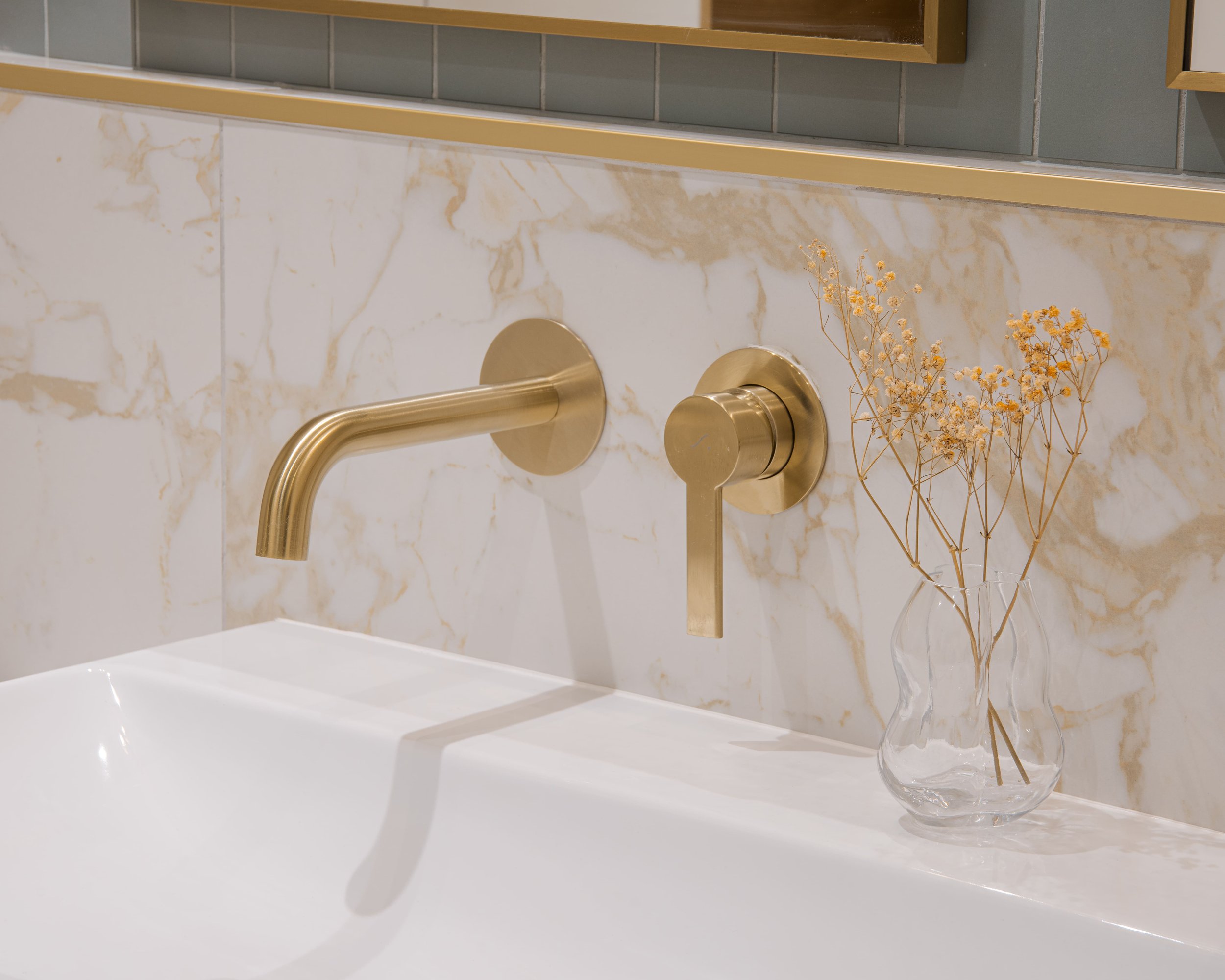 Lavabo blanc avec robinet en laiton, décor avec une vase en verre contenant des branches séchées dans une salle de bain moderne.