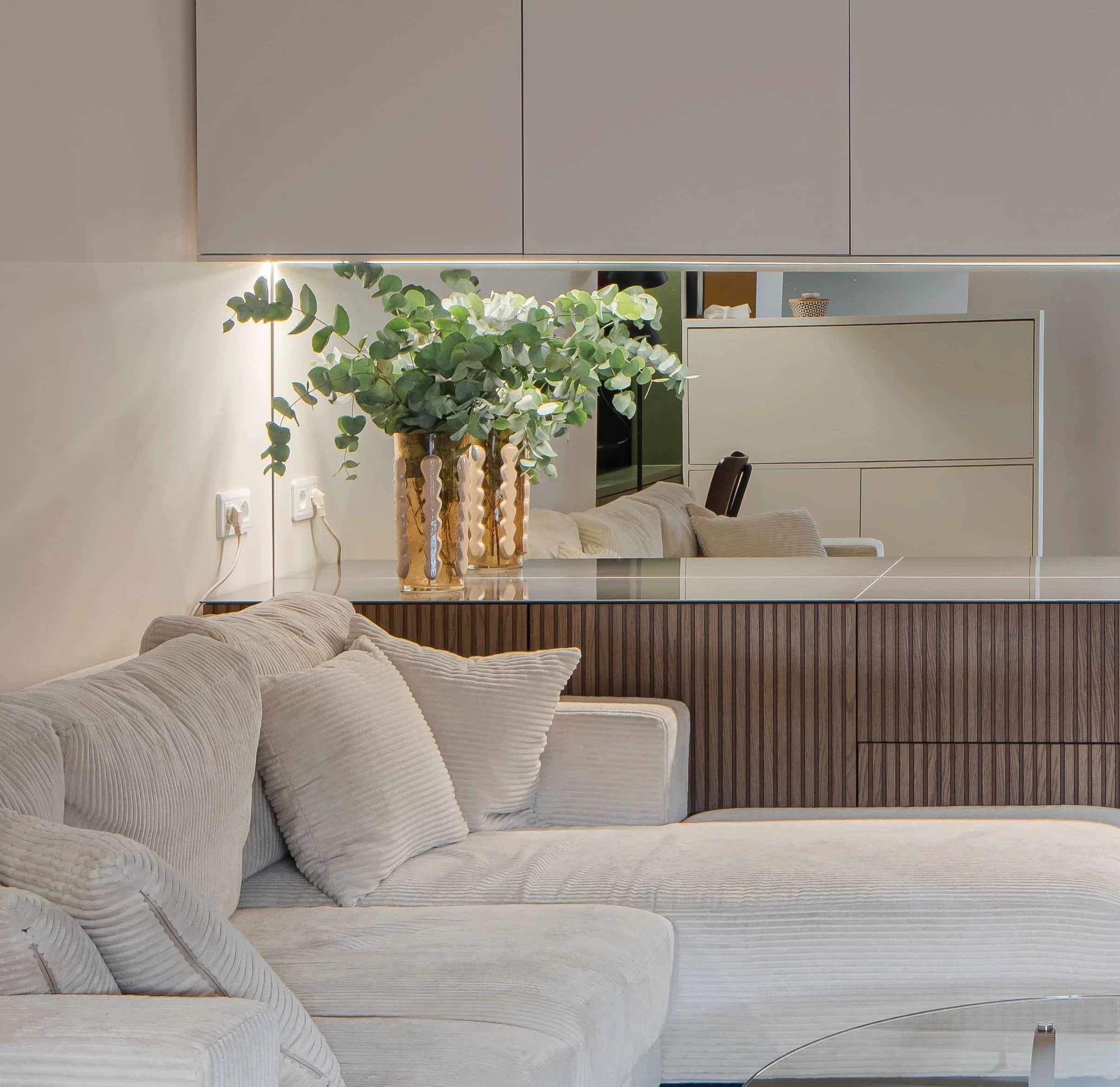Salon moderne avec canapé beige, coussins, grande plante dans un vase, et mobilier en bois avec miroir