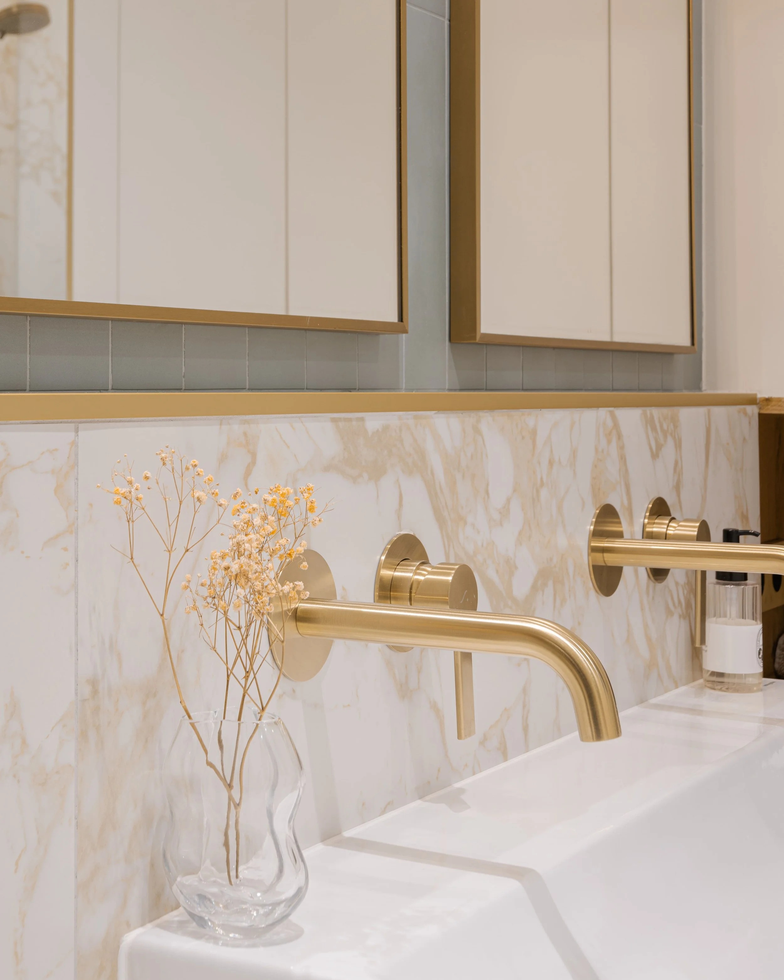 Lavabo avec un vase transparent contenant des bâtons de fleurs séchées, mural en marbre beige et blanc, miroir avec cadre doré, distributeurs de savon en laiton