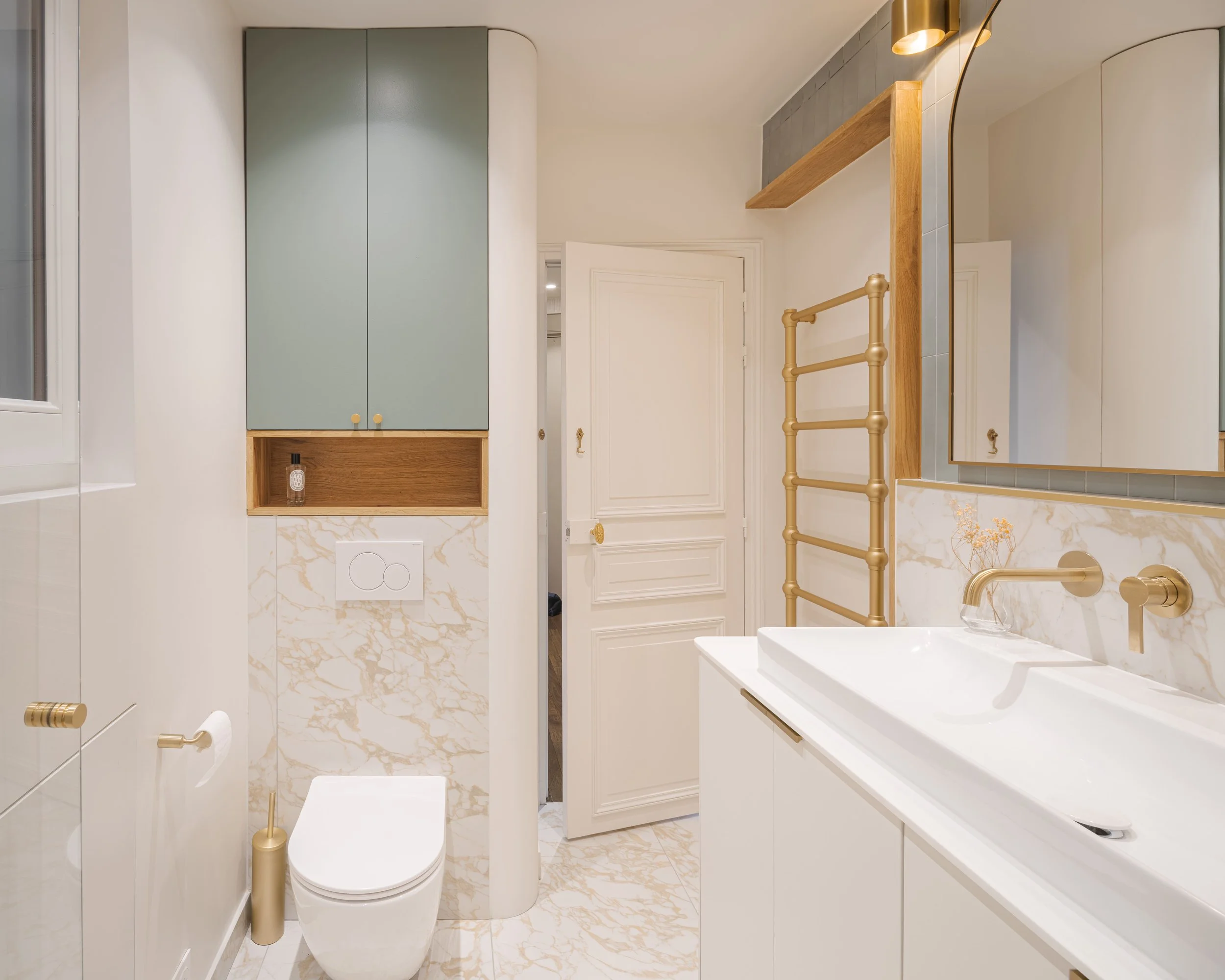 Salle de bain moderne avec lavabo blanc, miroir, meuble de rangement, toilette et accessoires dorés, murs en marbre et carrelage gris, porte blanche.