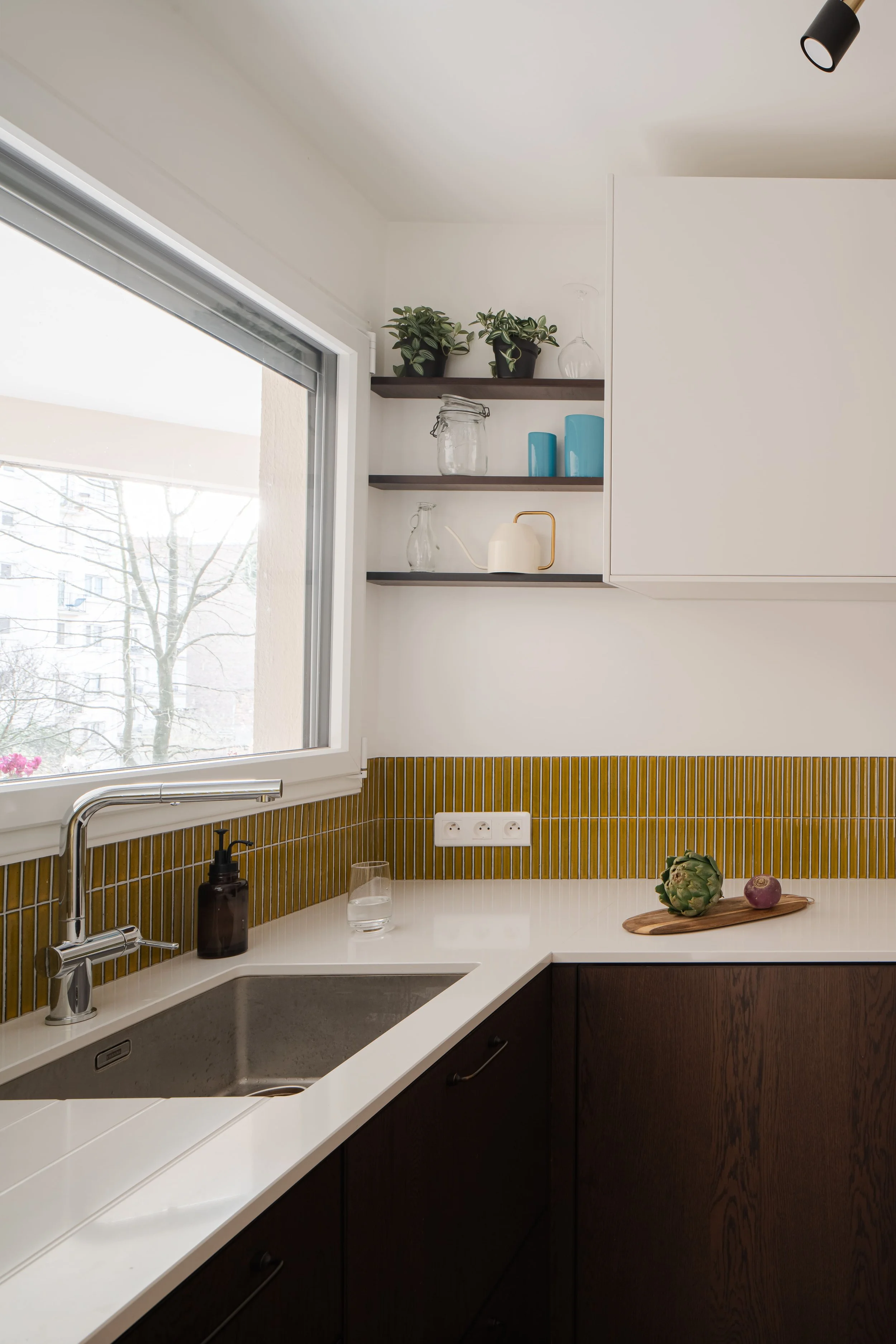 Cuisine moderne avec fenêtre, lavabo, étagères avec plantes et objets en verre, plans de travail en blanc et bois foncé, accents de couleurs vert et bleu.