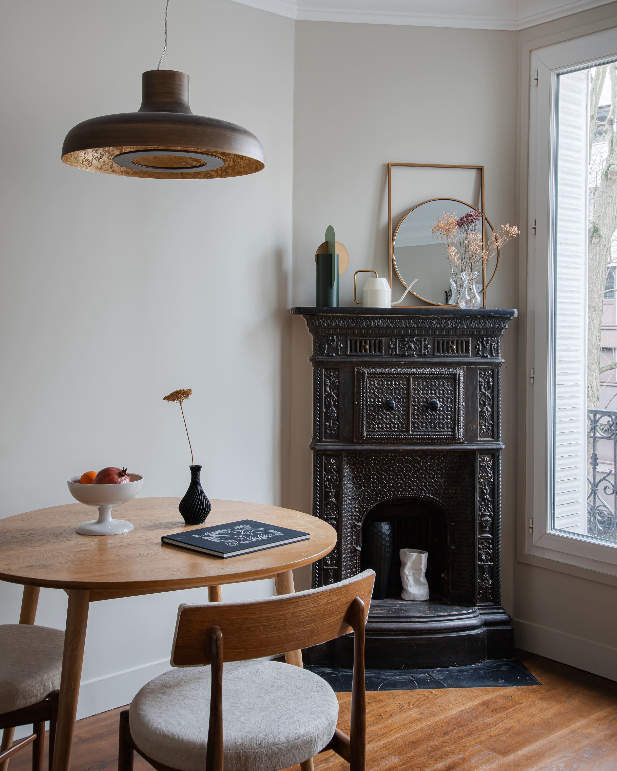 Intérieur d'une pièce avec un meuble en bois rond, deux chaises, un bol de fruits, et une cheminée noire ornée de décorations, près d'une grande fenêtre