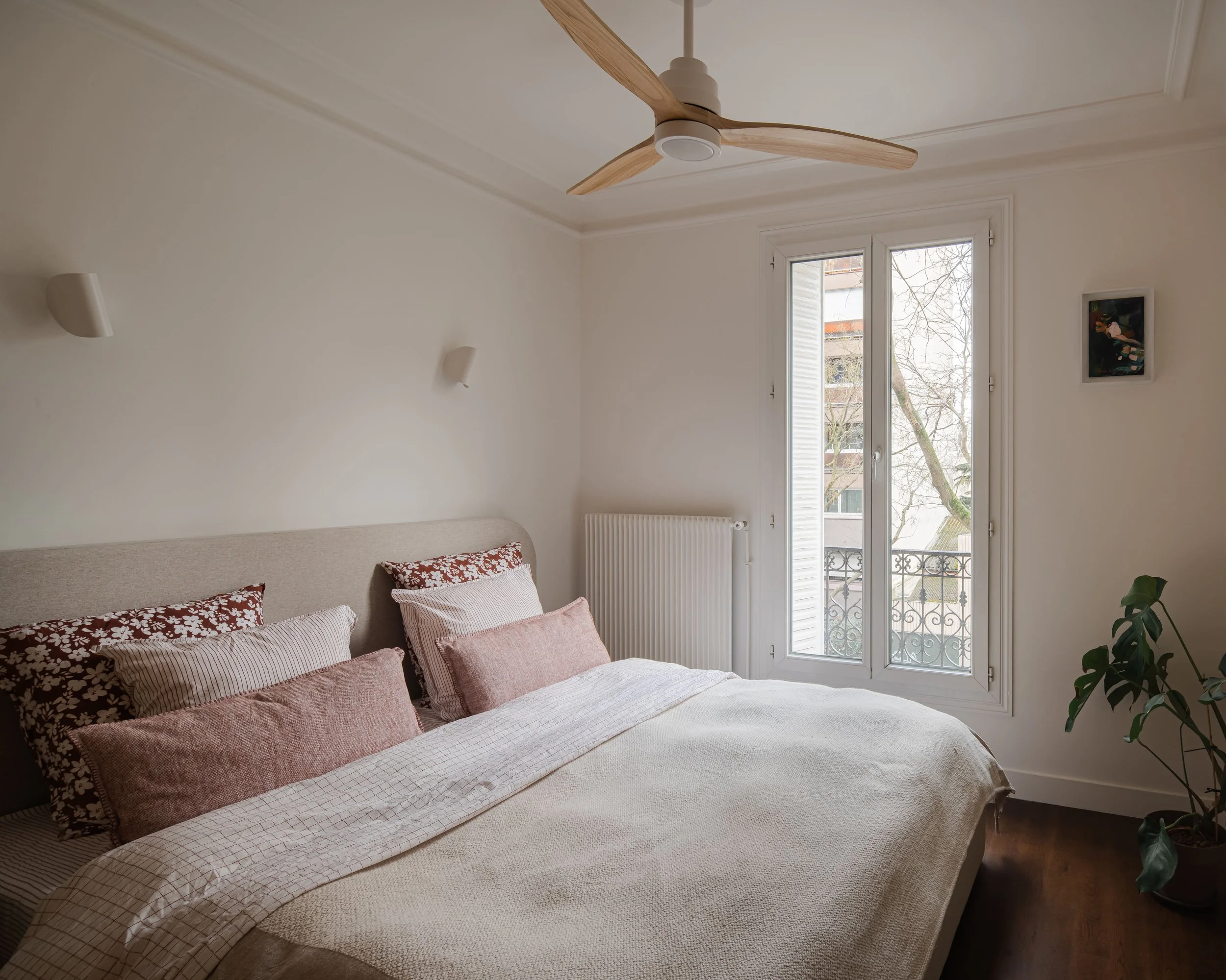 Chambre chaleureuse avec un lit fait, plusieurs coussins roses et à motifs floraux, une fenêtre laissant entrer la lumière naturelle, et une plante d'intérieur dans un pot en bas à droite.