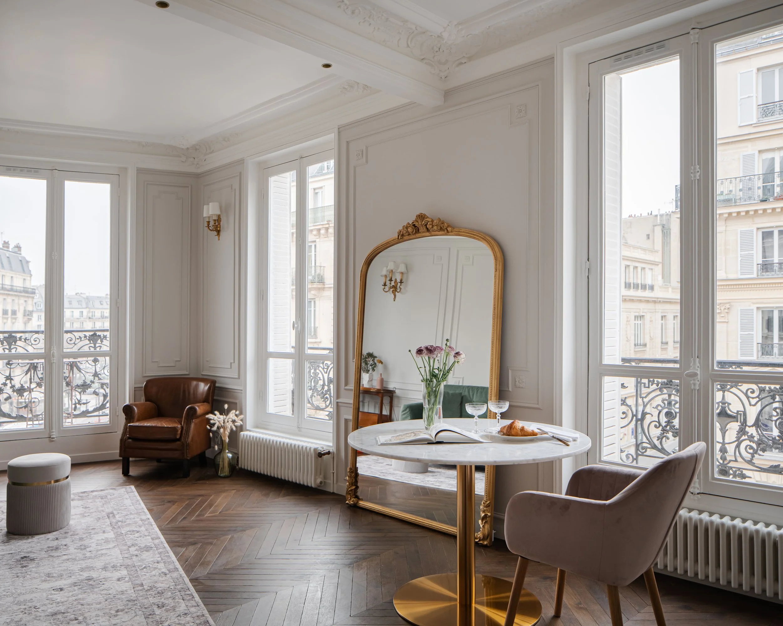 Salon avec fenêtres, miroir ancien, table ronde avec fleurs et viennoiseries, fauteuils, mur blanc décoré et parquet en bois.