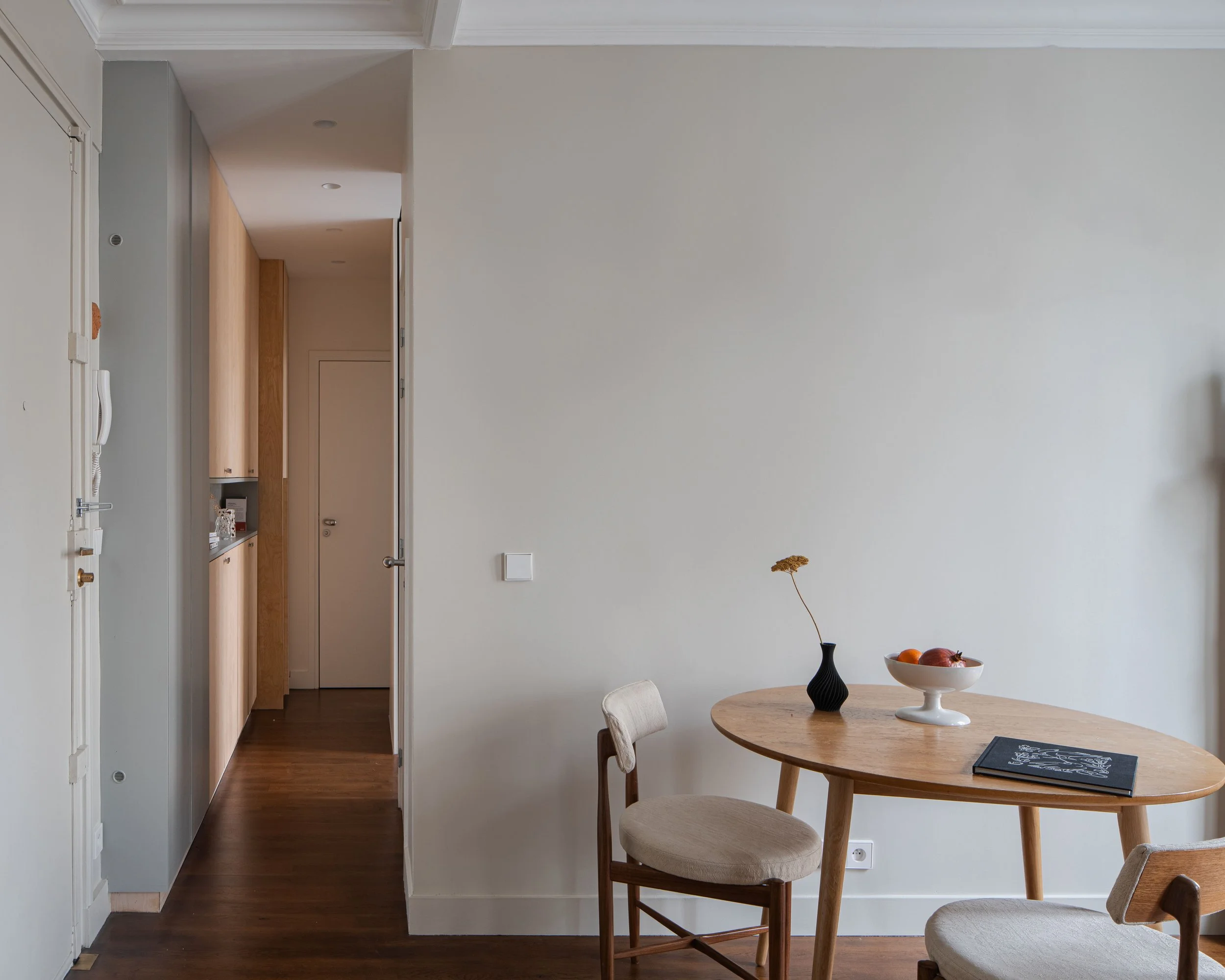 Intérieur d'une salle à manger claire avec table en bois, deux chaises beige, vase noir avec fleur, bol de fruits et un livre