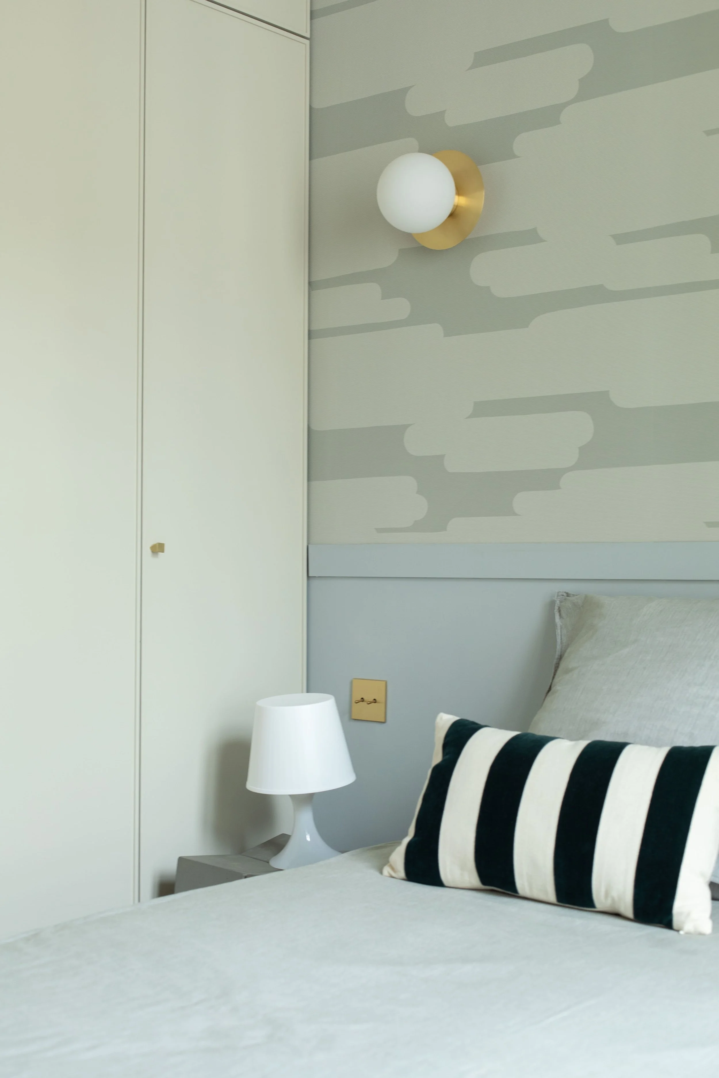Chambre moderne avec un lit, un coussin rayé noir et blanc, une lampe blanche sur une table de chevet, un mur avec un appliqué de lumière et une armoire