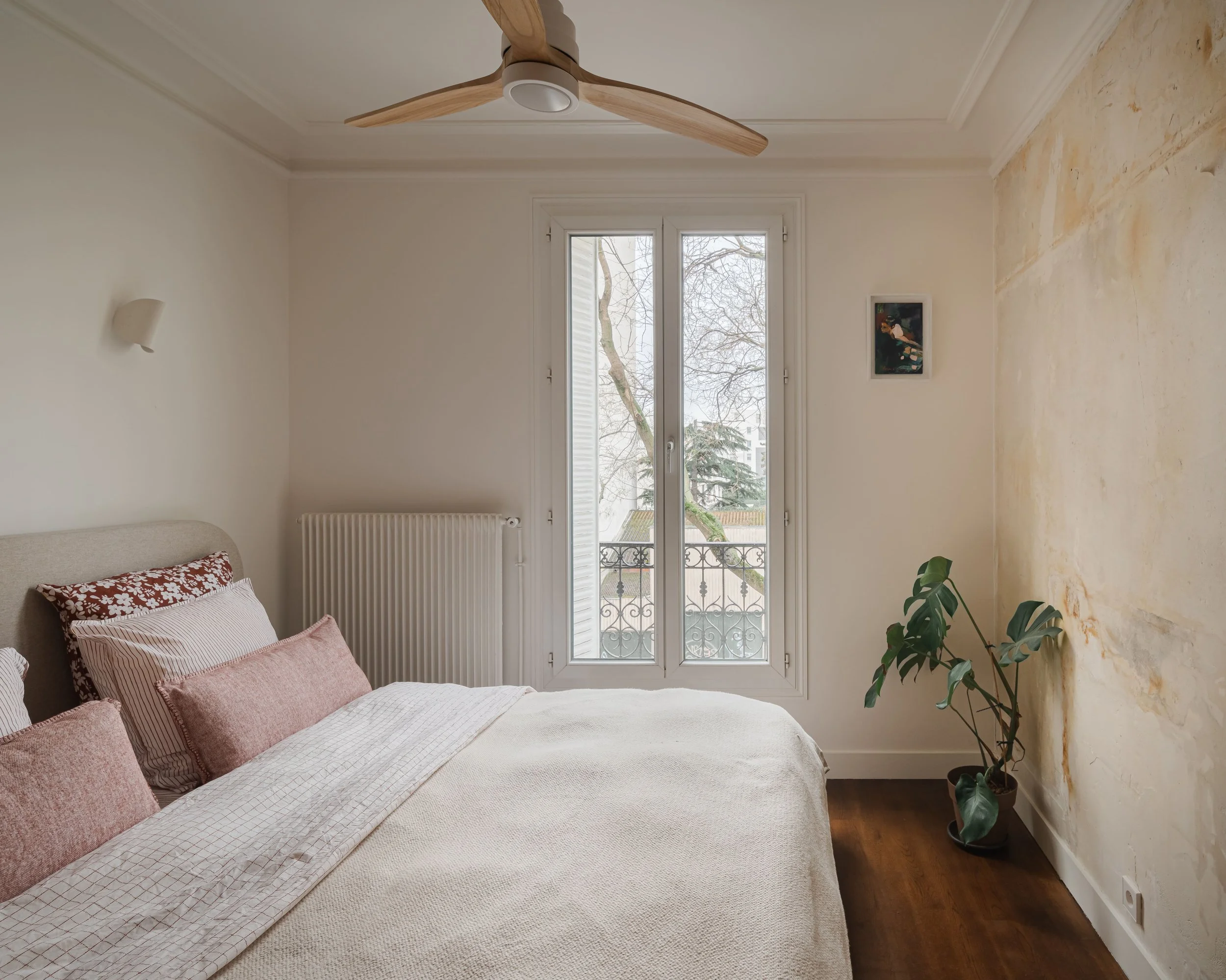Chambre minimaliste avec un lit, plusieurs oreillers, une fenêtre donnant sur un balcon avec vue sur des arbres, un mur bisé, une plante verte à droite, un cadre suspendu, une lampe murale et un ventilateur au plafond.