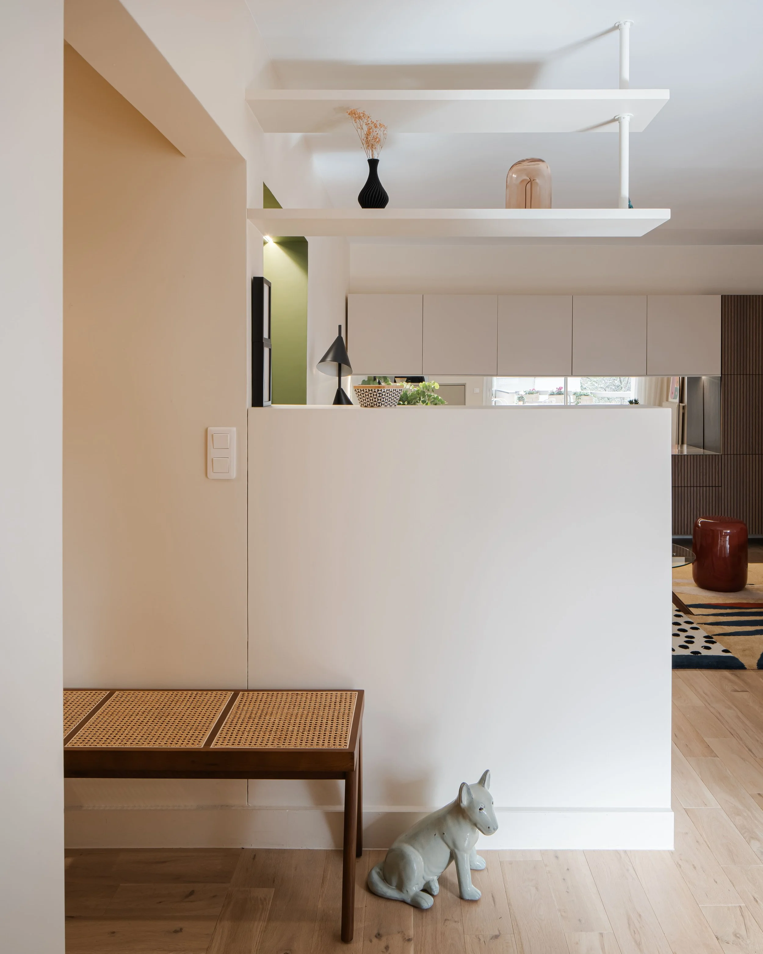Un coin intérieur d'une maison moderne avec un mur blanc, une petite sculpture de chien, un banc en bois, et des étagères suspendues avec des décorations. On voit aussi une partie de la cuisine en arrière-plan.