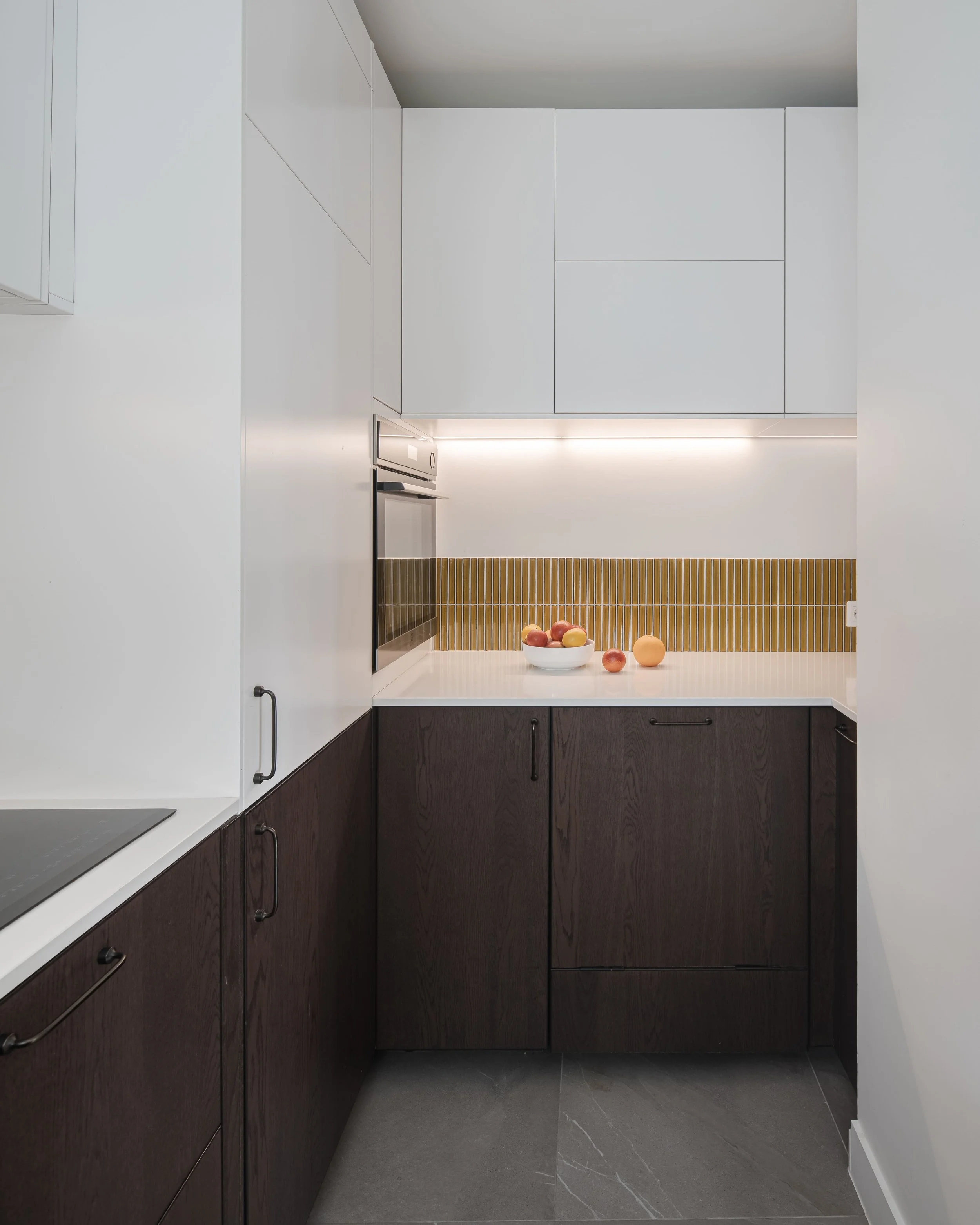 Petite cuisine moderne avec des armoires blanches en haut, des armoires en bois foncé en bas, un micro-ondes encastré et un bol de fruits sur le plan de travail.