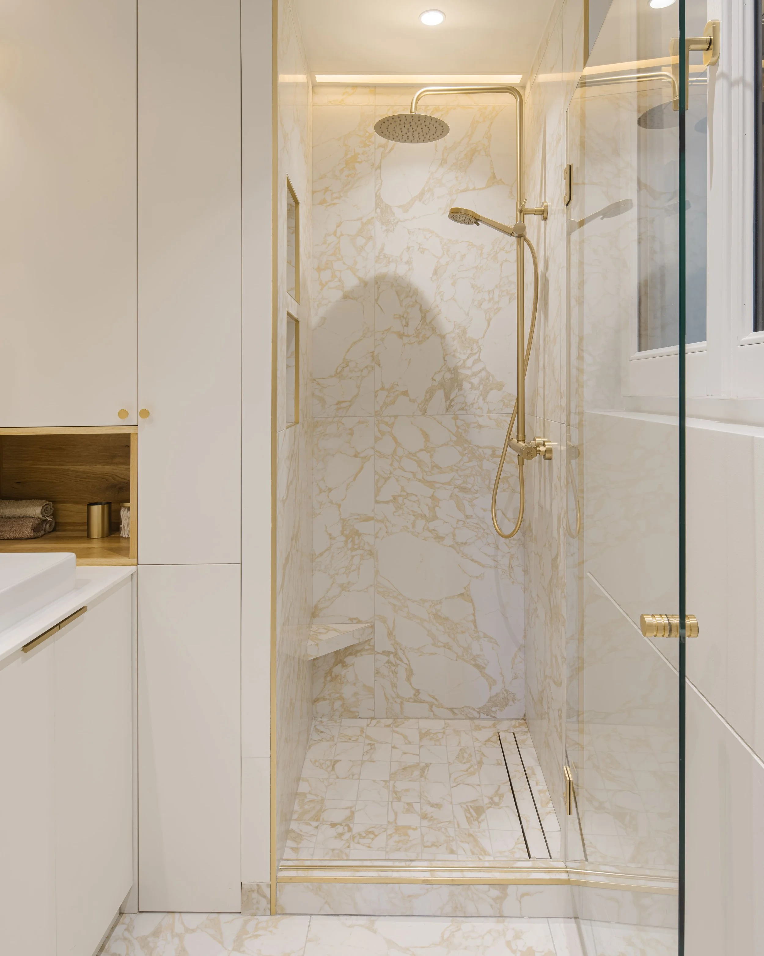 Douche en marbre blanc avec détails dorés et cabane en verre, avec fenêtre à droite.
