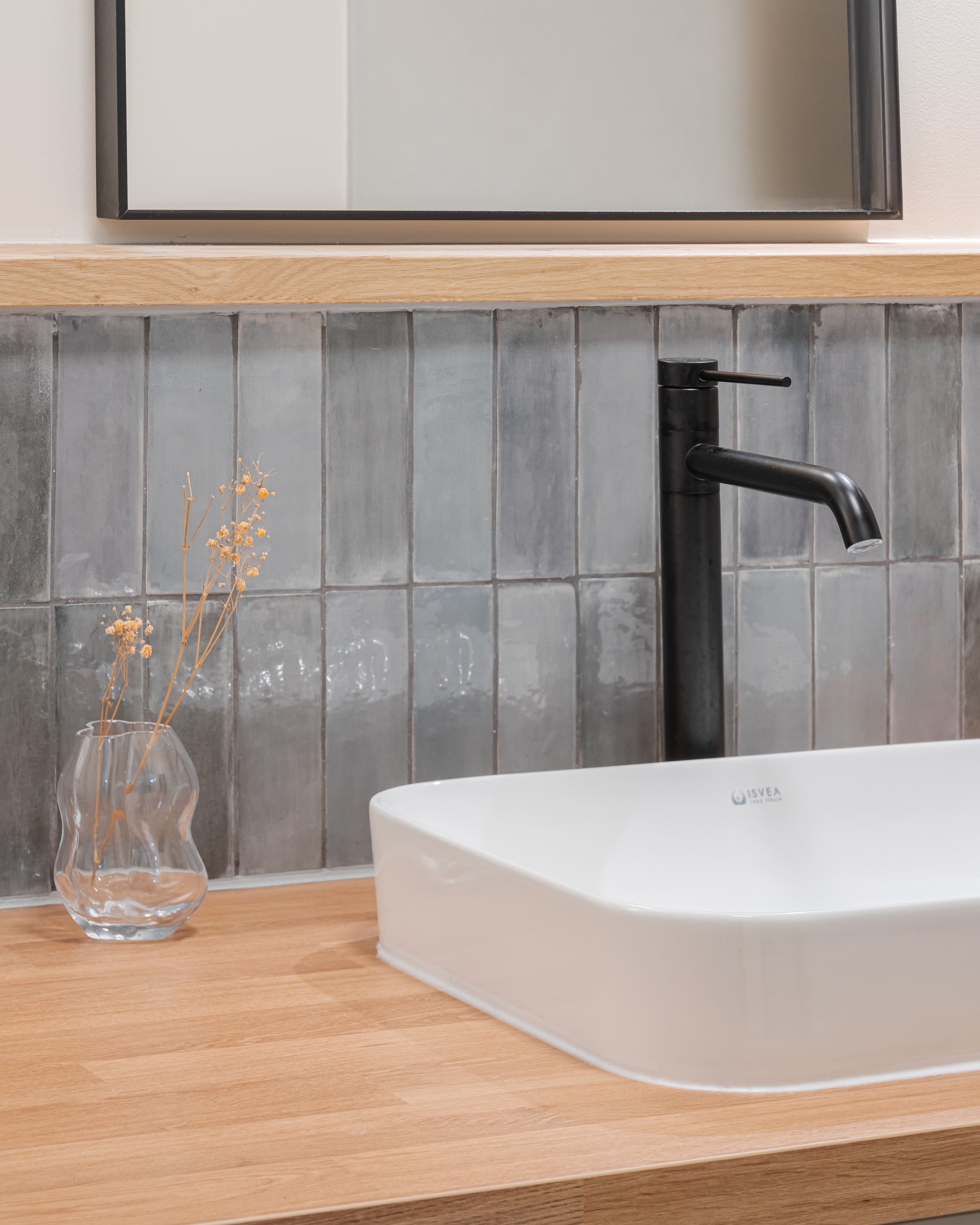 Lavabo blanc avec robinet noir, miroir au-dessus, vase en verre avec des branches sèches sur un plan de travail en bois, carrelage gris en fond.